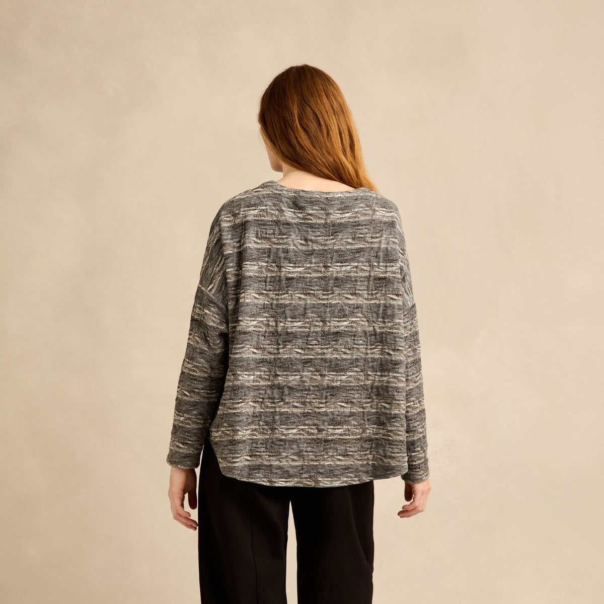 A woman is shown from behind wearing a gray and black patterned knit sweater, black pants, and black boots against a neutral beige studio backdrop.