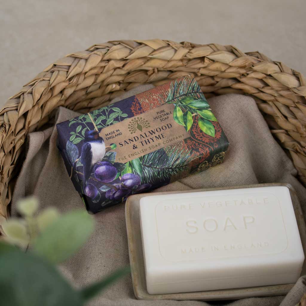 A bar of soap in a clear plastic container placed beside its illustrated wrapper inside a woven straw basket on a beige fabric surface.