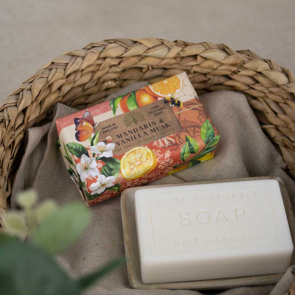 A bar of soap in a clear plastic tray sits beside its colorful illustrated wrapper inside a woven straw basket lined with beige fabric. The wrapper reads 'Mandarin & Vanilla Musk' and shows citrus, flowers, and a butterfly.