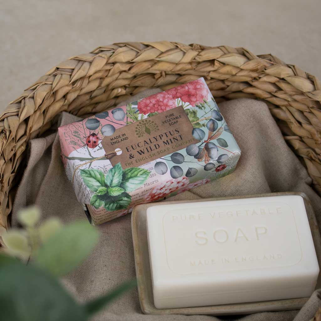 Two bars of soap displayed in a woven straw basket lined with beige fabric. One bar is wrapped in a pink botanical paper sleeve labeled 'Eucalyptus & Wild Mint' by Pure Vegetable, and the other is a plain off-white bar embossed with 'PURE VEGETABLE SOAP'.