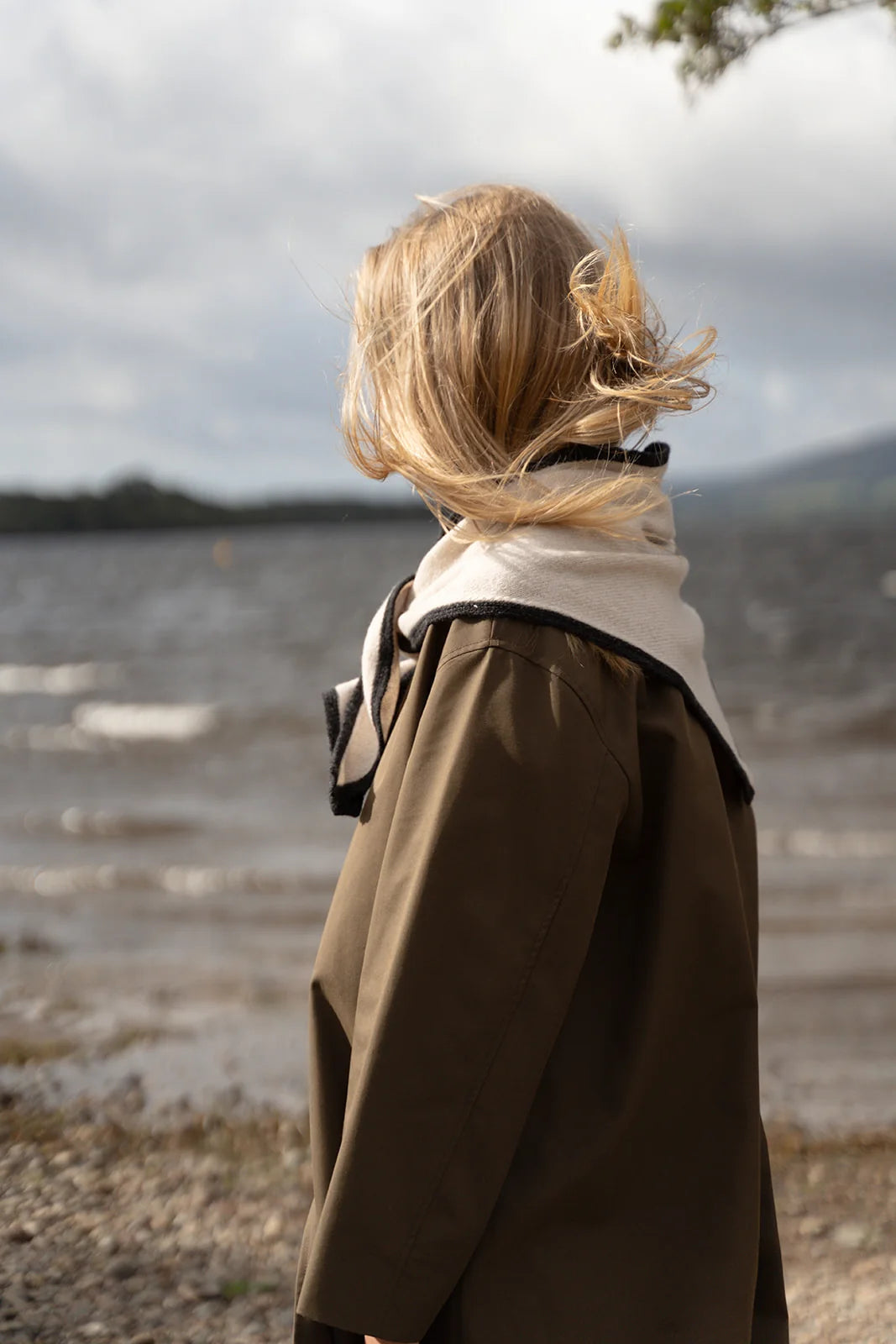 A child seen from behind wearing a brown coat with a cream collar, standing outdoors near a body of water under an overcast sky.
