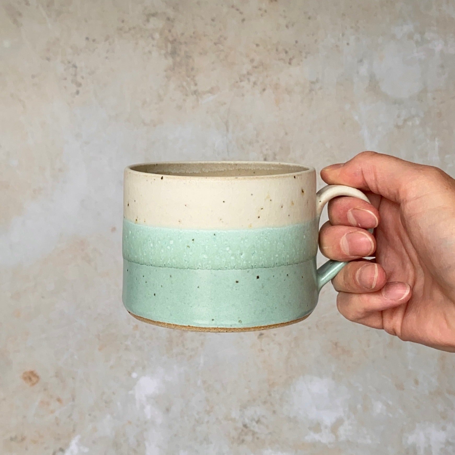 A speckled, two-toned ceramic mug; off-white top and light teal bottom, held in a hand.