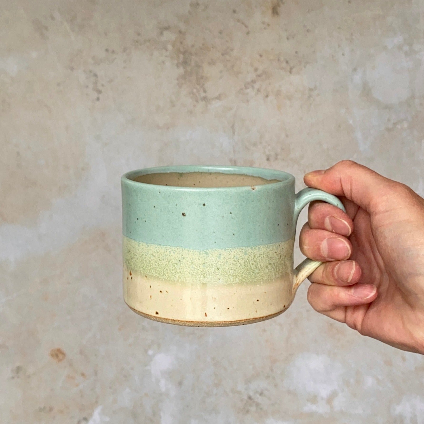 Hand holding a speckled, light-blue, cream, and green ceramic mug with a rounded handle.