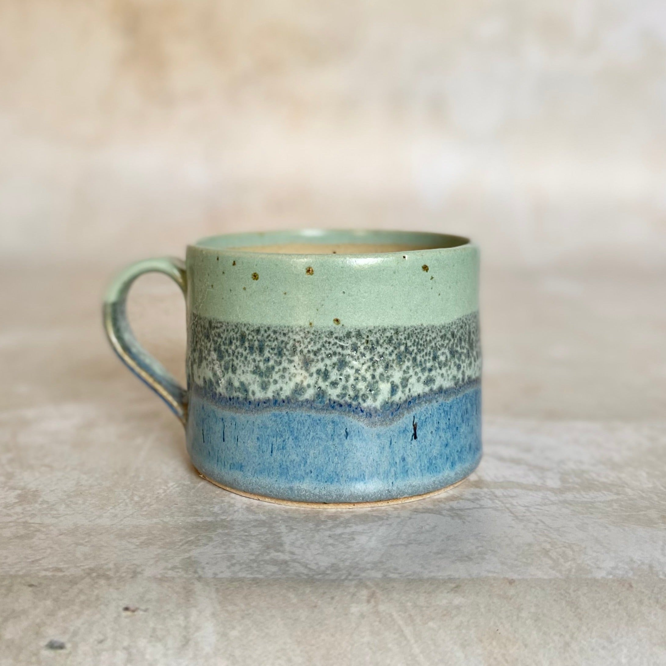 Hand-thrown ceramic mug with a light green, speckled gray, and blue glaze in horizontal bands.