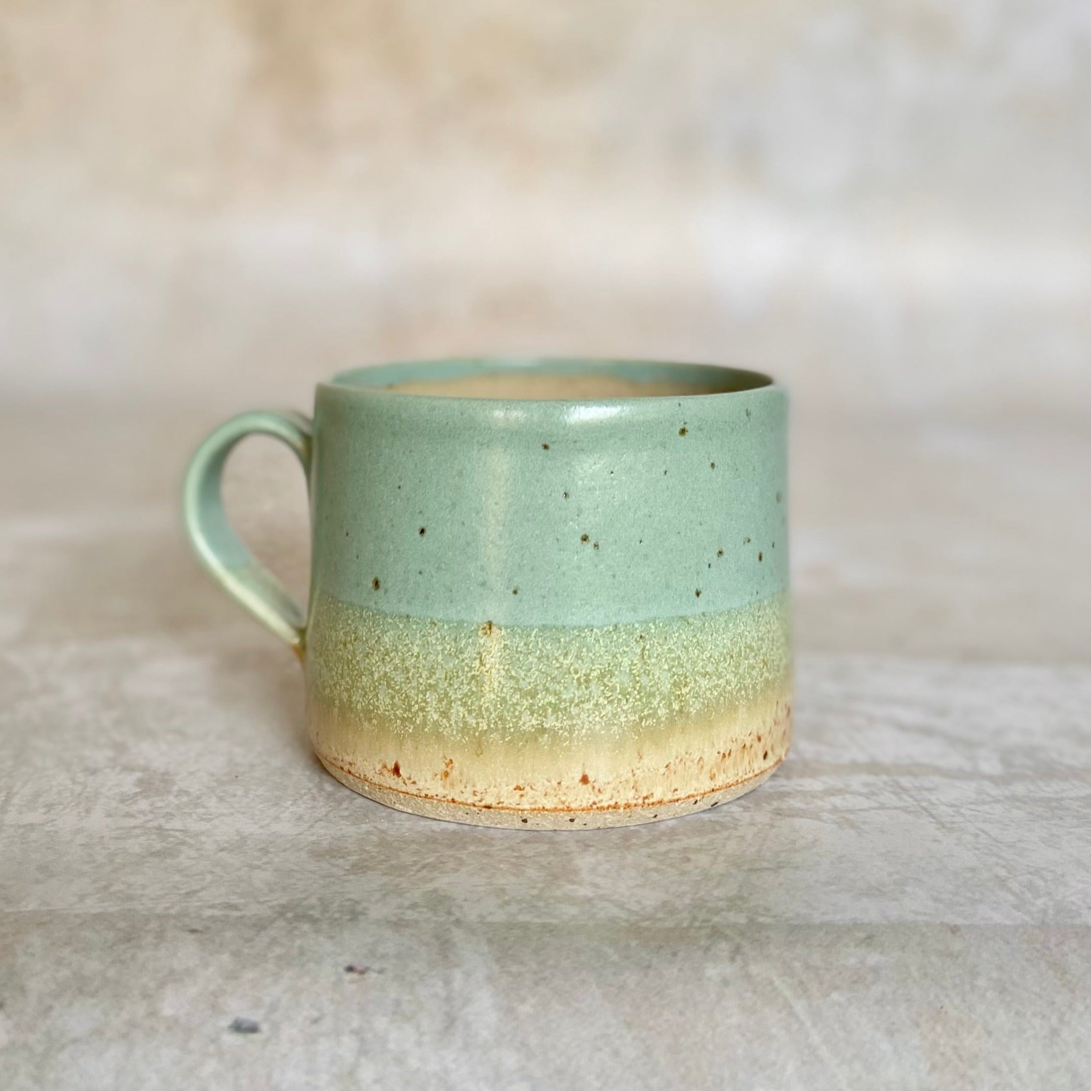 A speckled light-green and beige ceramic mug with a rounded handle.  The mug has a subtle two-toned glaze.