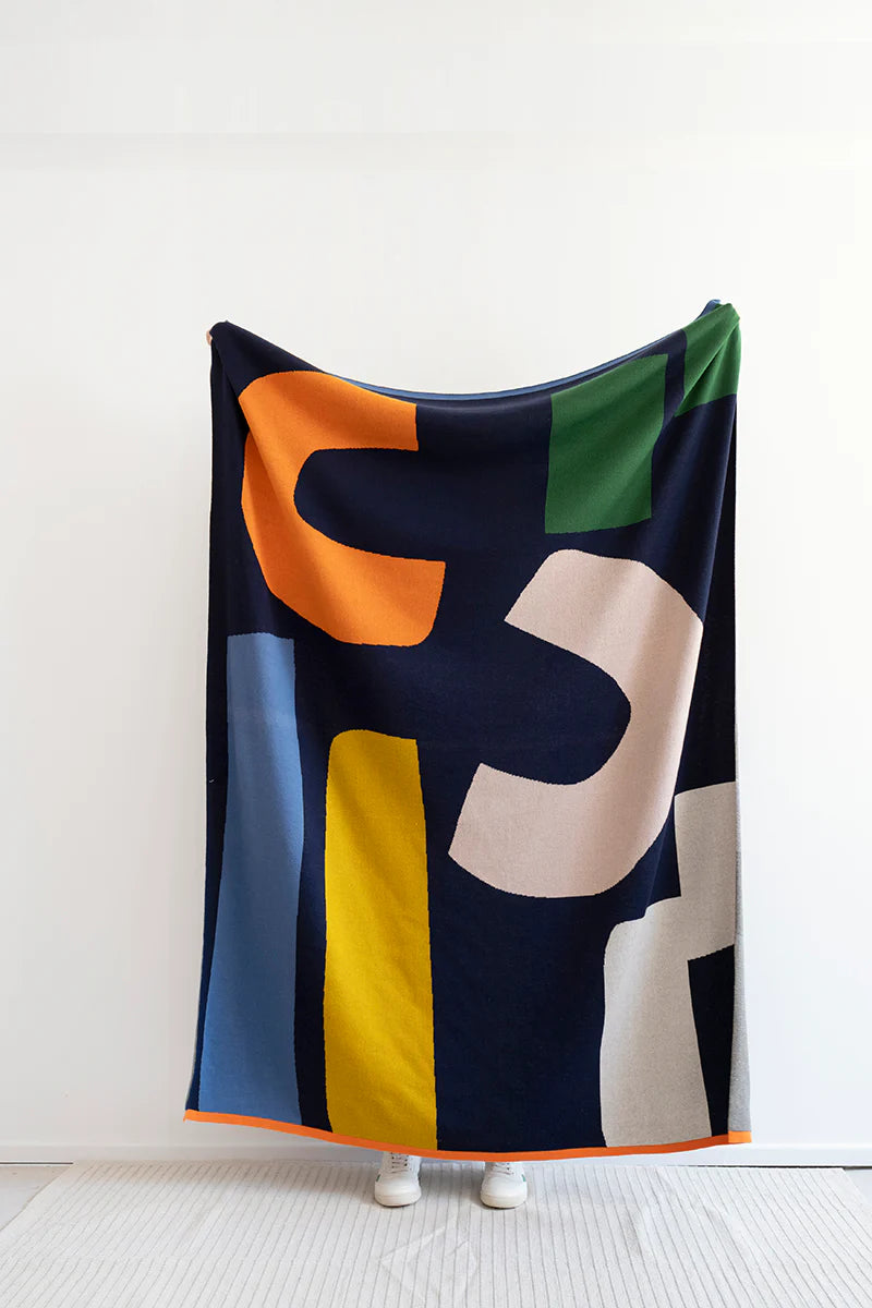 Large knit throw blanket in navy blue with bold, colorful abstract shapes in orange, yellow, light beige, and green.