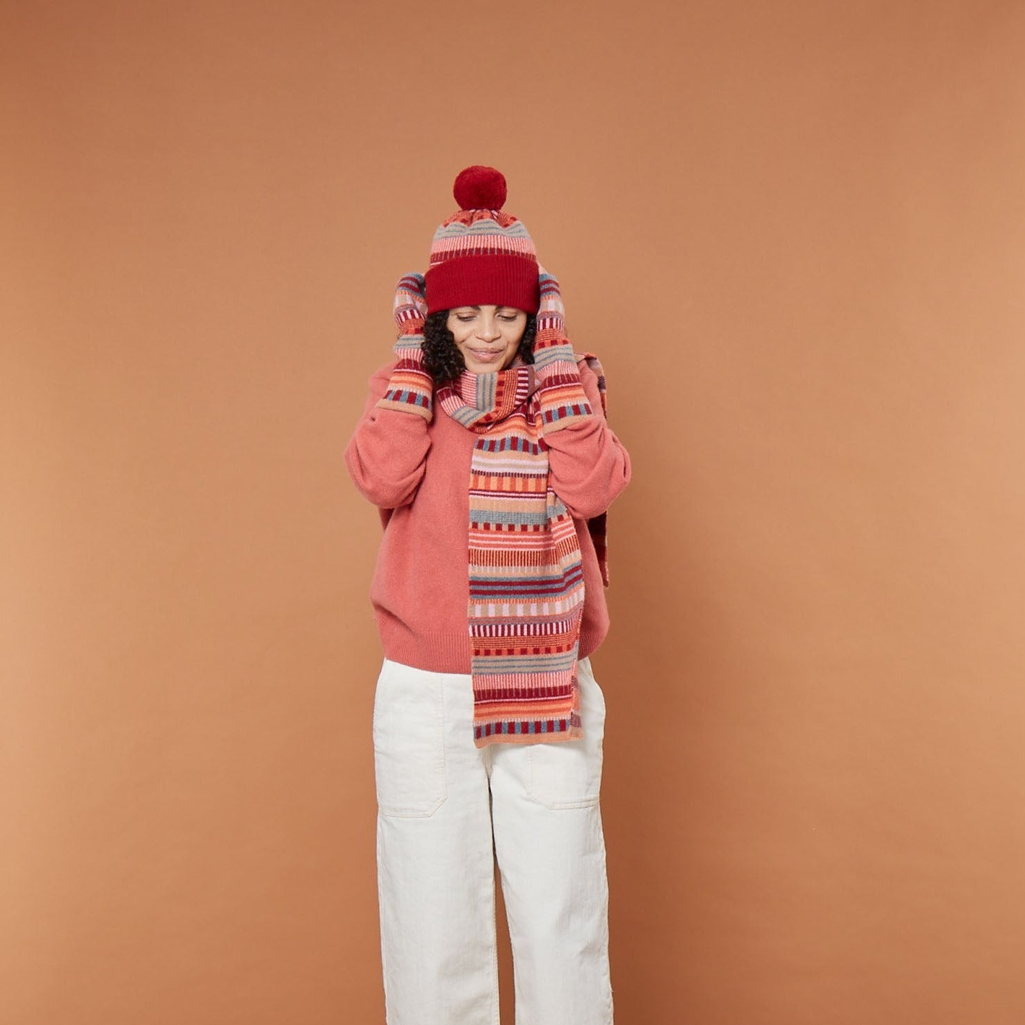 A woman in a pink sweater, red hat, and patterned scarf from Donna Wilson, posing against a brown background.