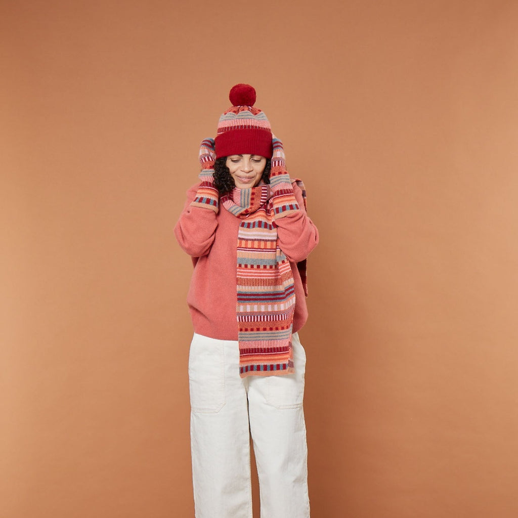 A woman in a pink sweater, red hat, and patterned scarf from Donna Wilson, posing against a brown background.