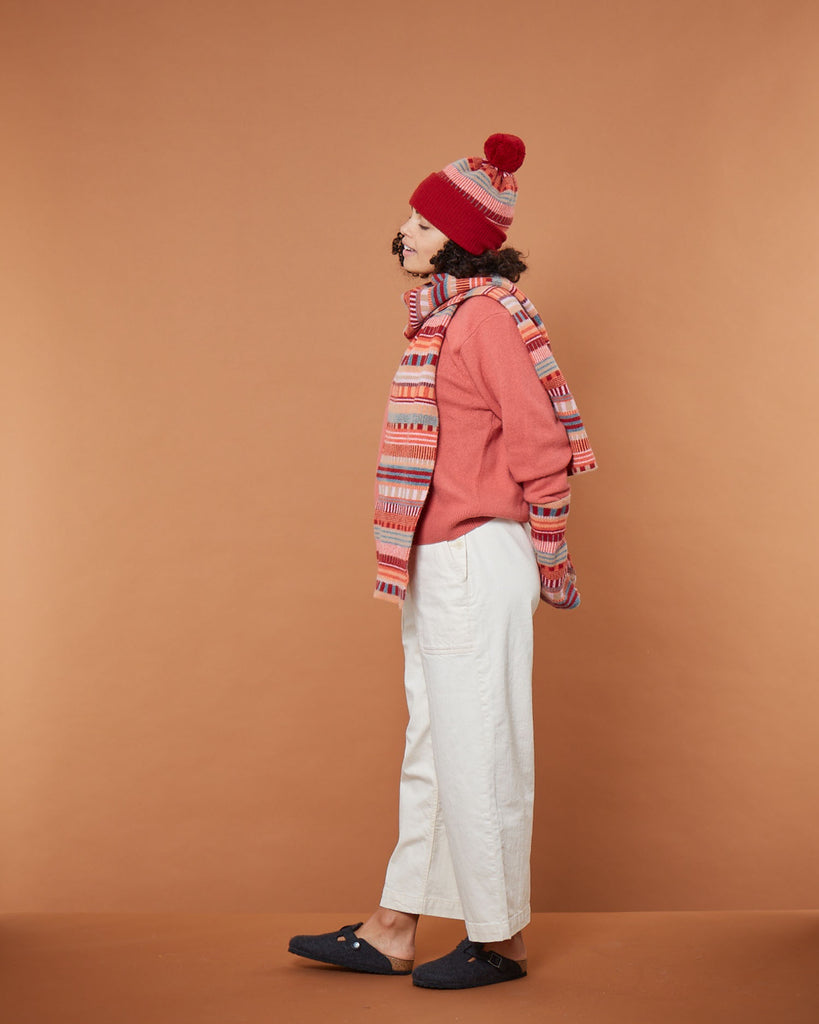 Woman in a pink sweater, red hat, patterned scarf, white pants, and grey shoes by Donna Wilson, against a brown backdrop.