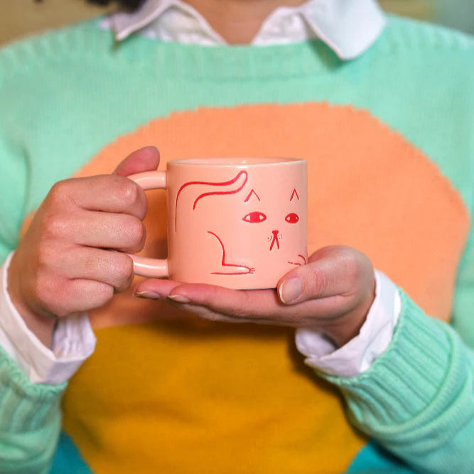 Person holding a pink mug with a cat illustration, wearing a colorful sweater.