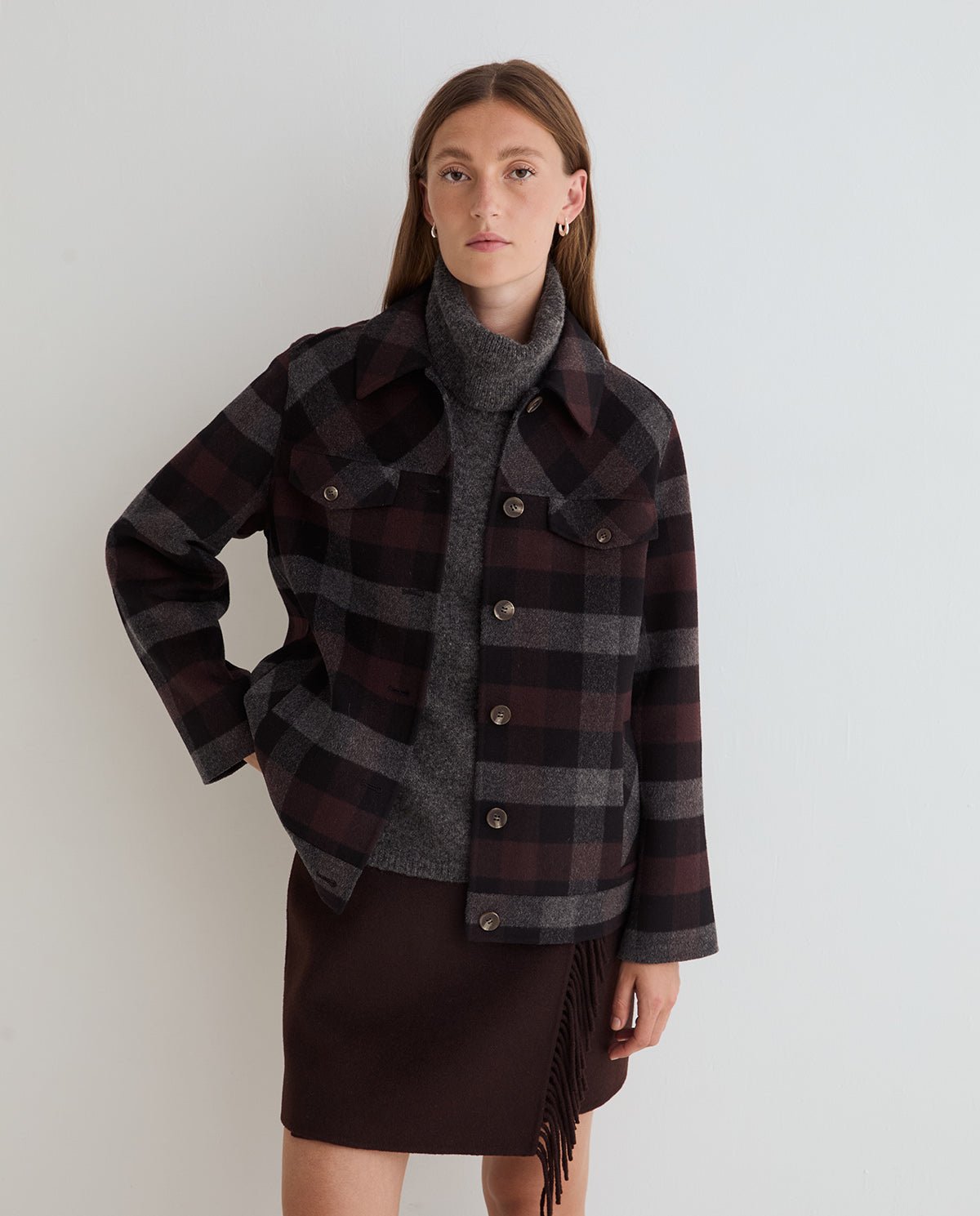 A model wearing a dark brown and gray plaid button-up jacket over a gray turtleneck sweater and a dark brown fringe skirt, standing against a light gray studio background.