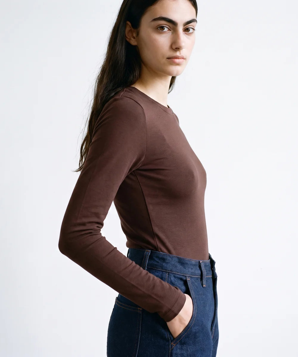 A woman wearing a fitted, long-sleeve crew-neck top in a dark brown color, paired with dark blue jeans, photographed against a clean white studio background.
