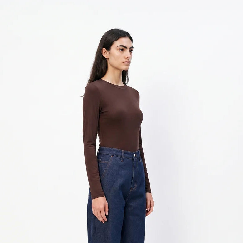 A woman stands in profile wearing a fitted brown long-sleeve top, dark blue wide-leg jeans, and black flats against a plain white studio background.