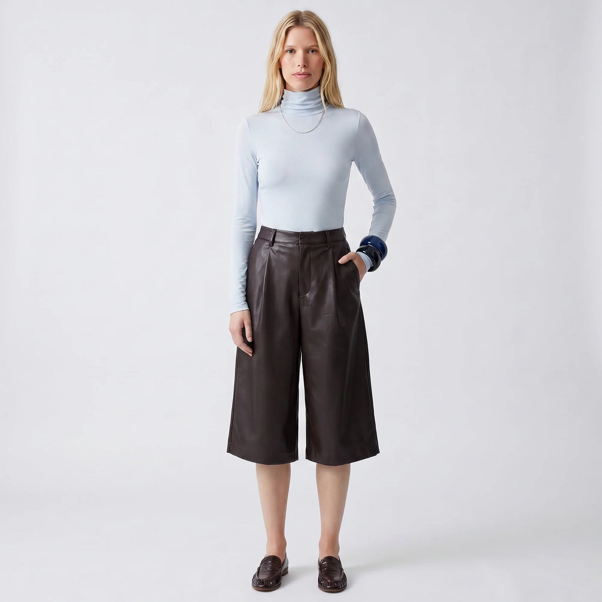 A woman stands against a light gray studio background wearing a light blue long-sleeve mock-neck top, dark brown wide-leg faux-leather cropped pants, and dark brown loafers.