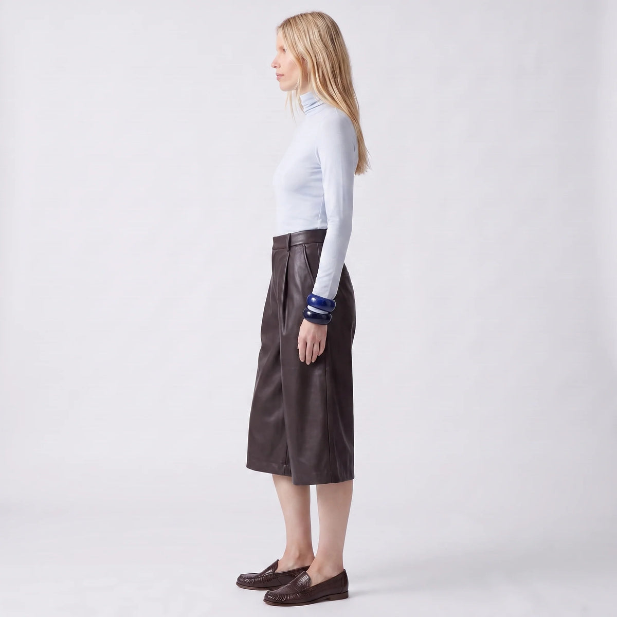 A woman stands in profile wearing a light blue long-sleeve mock-neck top, a dark brown leather midi skirt, and dark brown loafers against a plain light gray studio background.