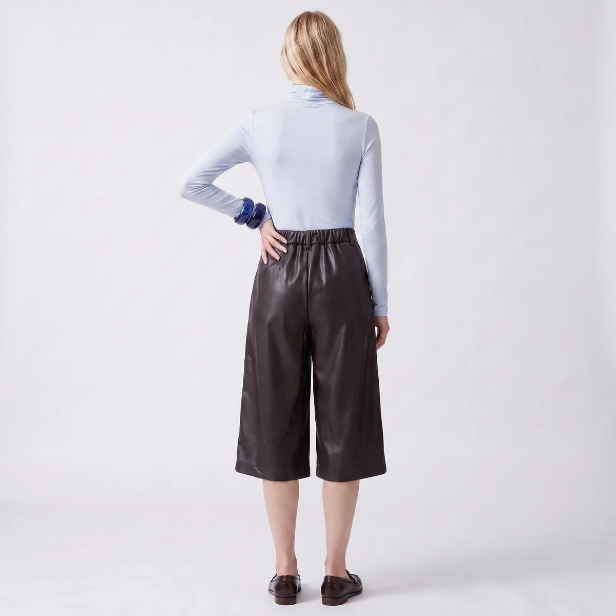 A woman shown from the back wearing a light blue long-sleeve mock-neck top and dark brown faux-leather cropped pants, paired with dark brown loafers, against a light gray studio background.