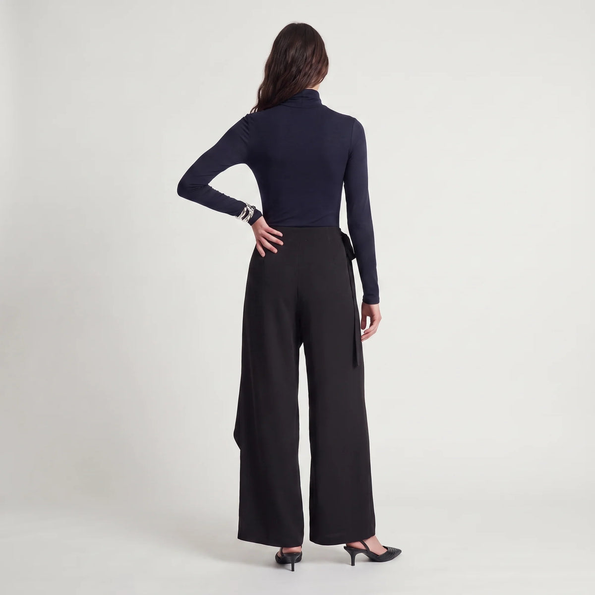 A woman is shown from the back wearing a navy blue long-sleeve mock neck top and black wide-leg trousers, paired with black heels, against a plain light gray studio background.