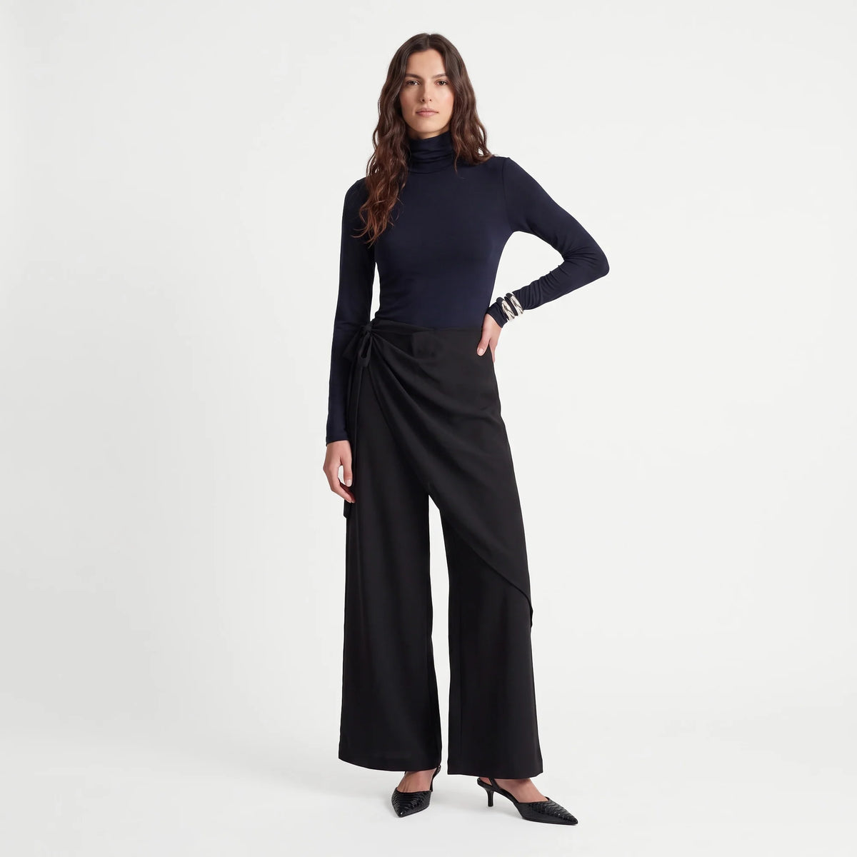 A woman modeling a dark navy long-sleeve mock neck top paired with black wide-leg draped pants and black pointed-toe heels, photographed against a clean light gray studio background.
