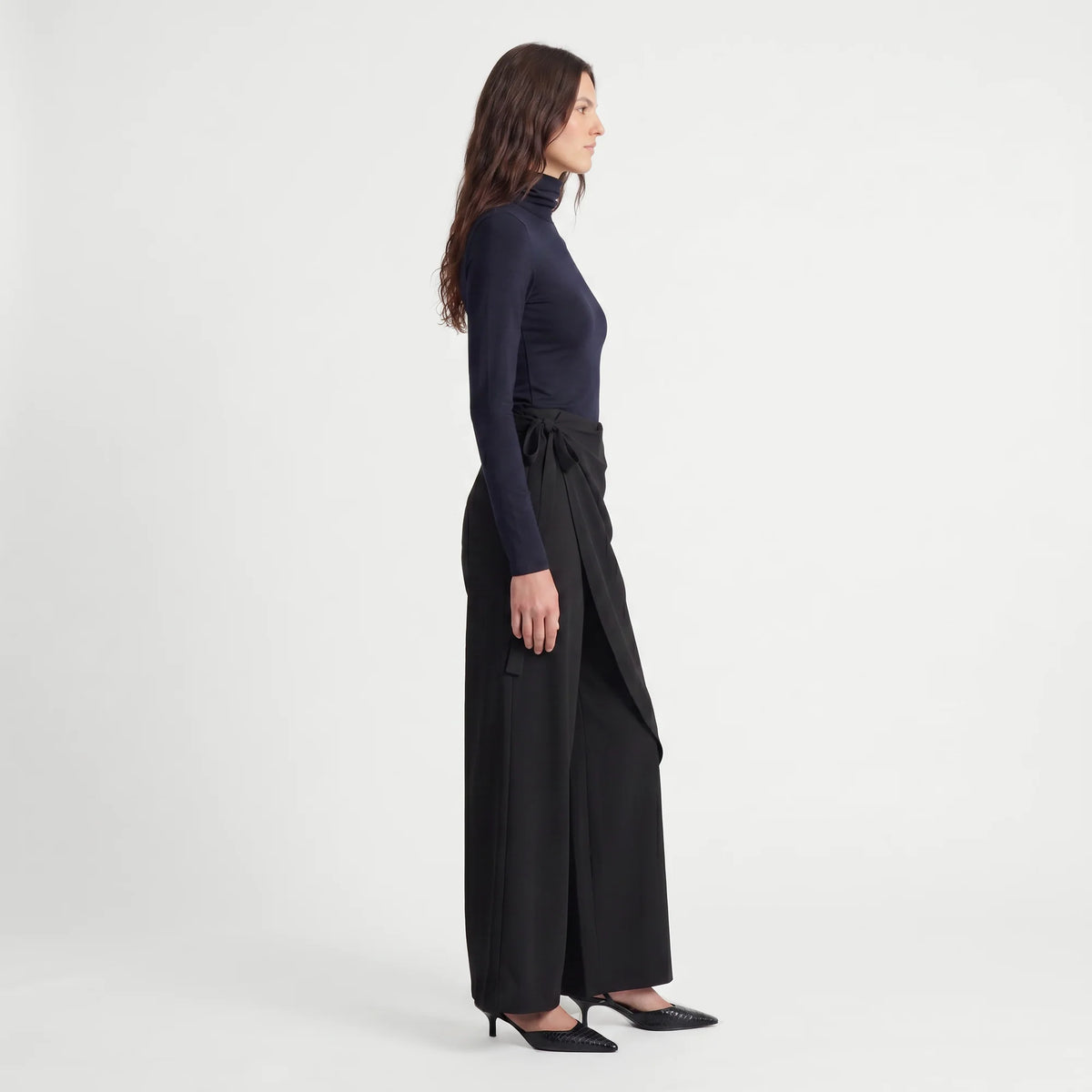 A woman in side profile wearing a dark navy mock-neck long-sleeve top and black wide-leg trousers with a side tie, paired with black pointed-toe heels, against a plain light gray studio background.