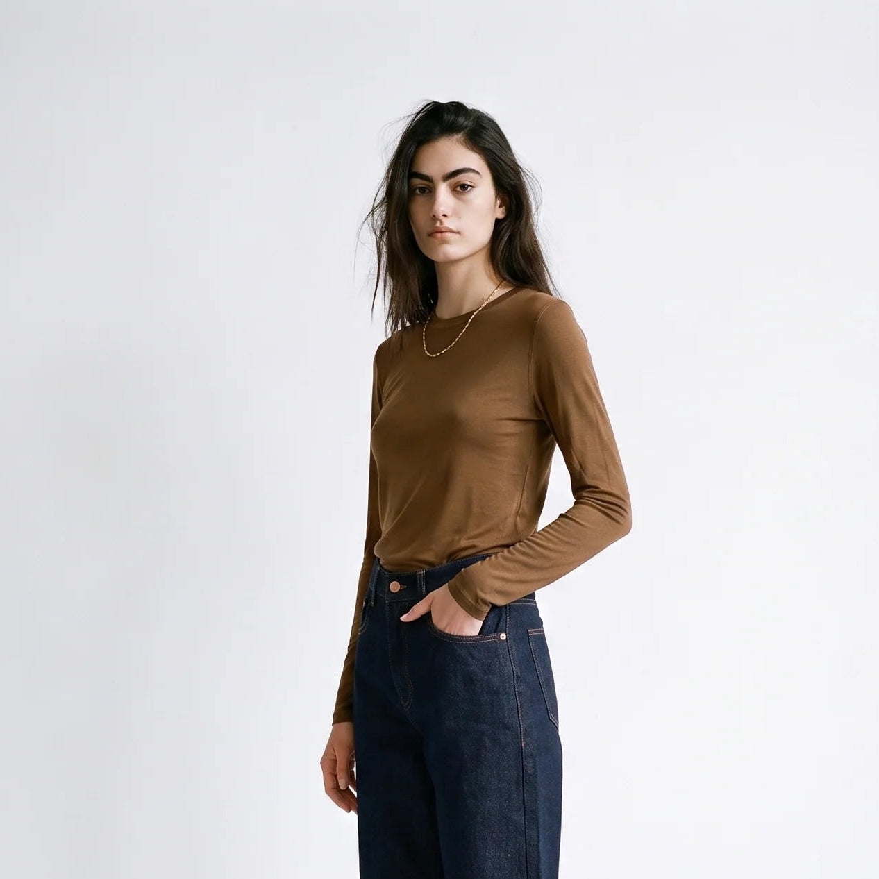 A woman modeling a brown long-sleeve knit top and dark blue wide-leg jeans with black mule shoes, standing against a plain white studio background.