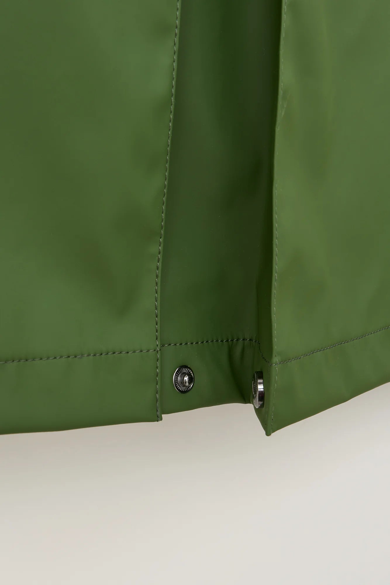 Close-up of an olive green jacket or raincoat showing the side seam, button snap closure, and fabric texture against a light neutral background.