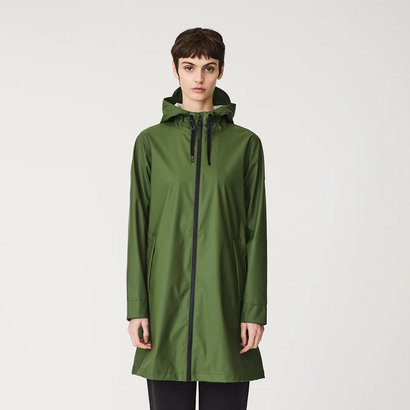 A model stands against a light gray studio background wearing a forest green hooded raincoat with a black front zipper, black pants, and hiking boots.