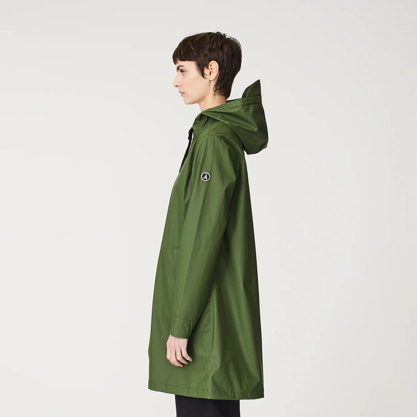 A model shown in profile wearing a mid-length olive green hooded raincoat, black pants, and hiking boots against a plain light gray studio background.