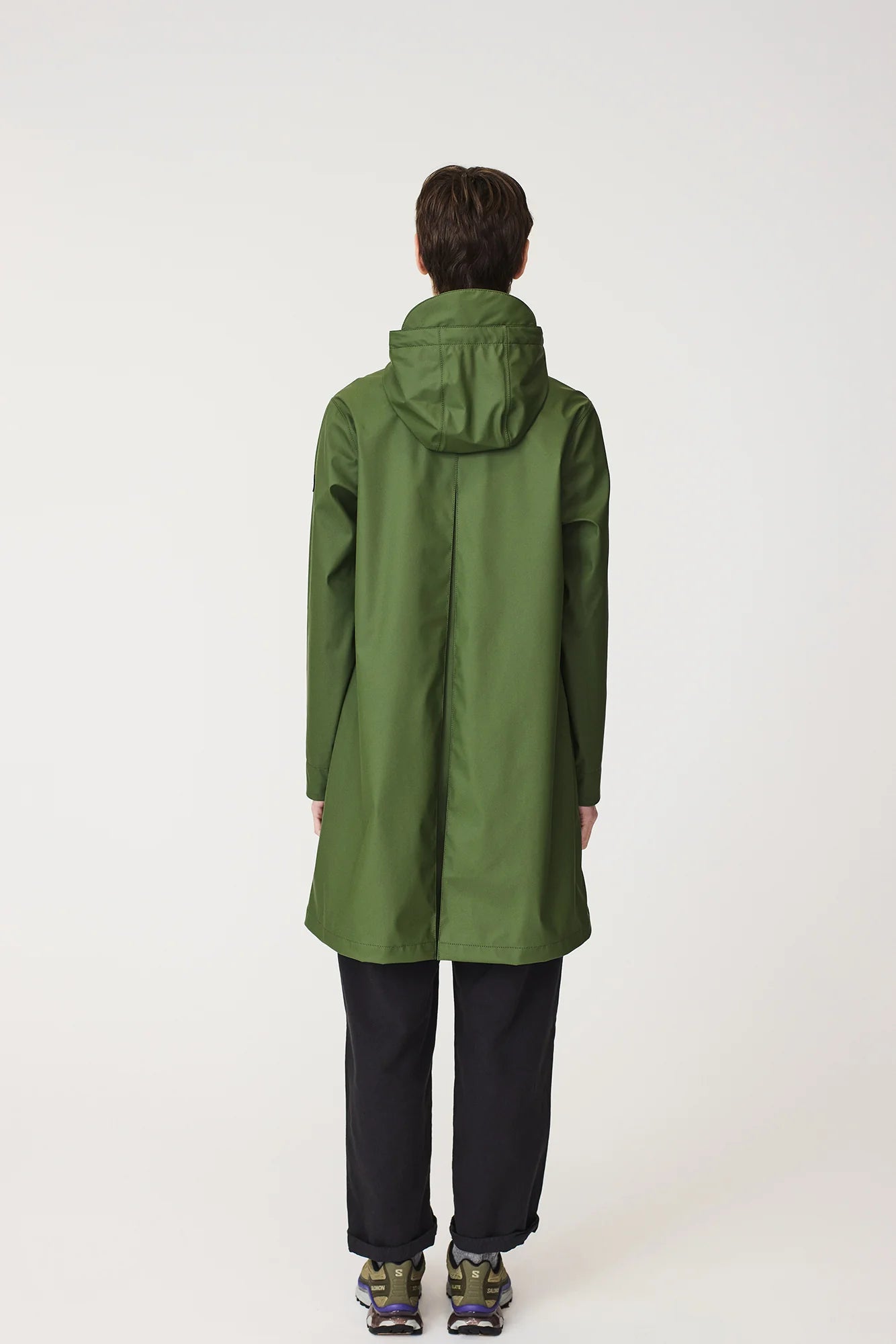 A model viewed from the back wearing a mid-length olive green hooded raincoat, black trousers, and hiking boots against a plain white studio background.