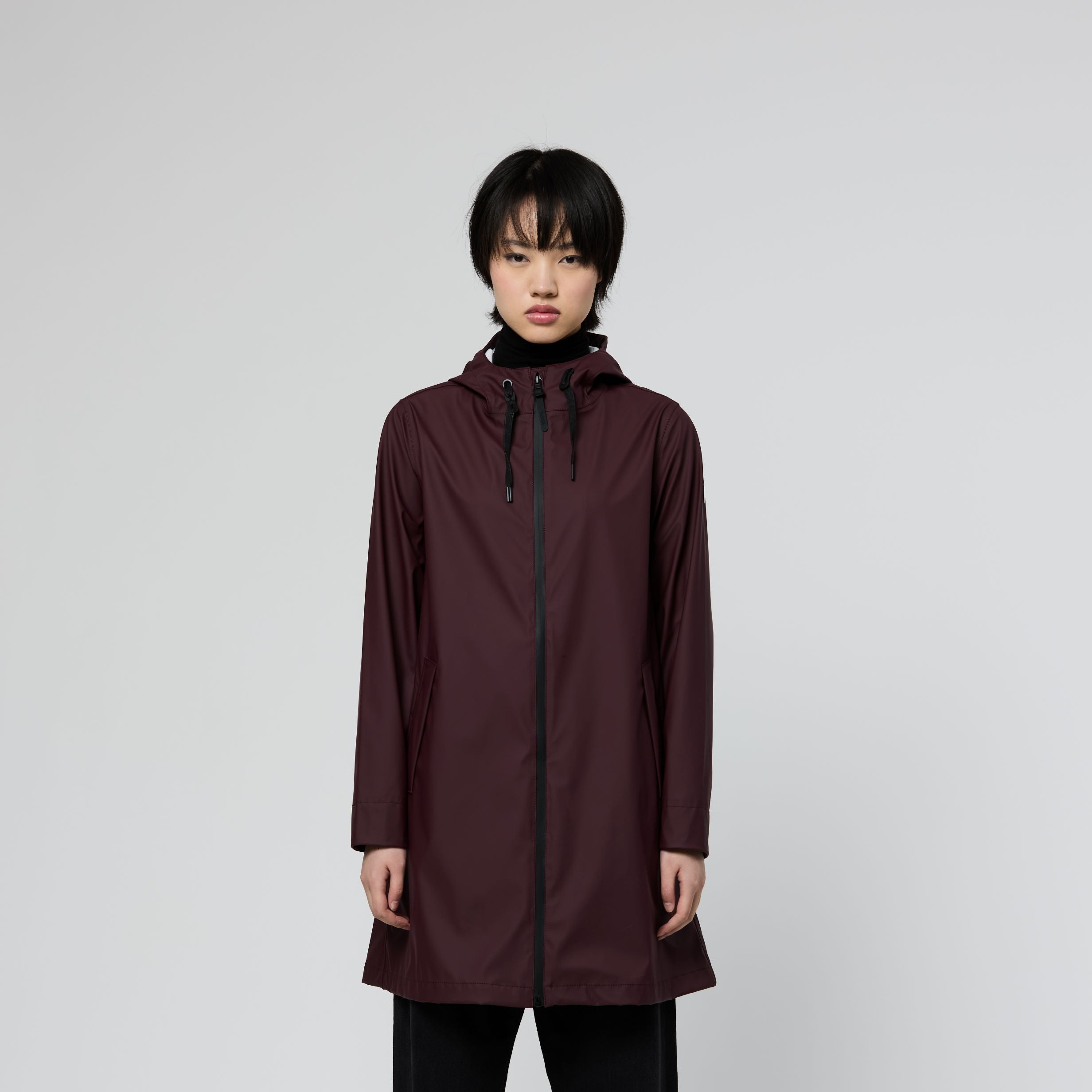A model stands against a light gray studio background wearing a long, dark burgundy hooded raincoat with black drawstrings, black wide-leg pants, and black-and-white sneakers.