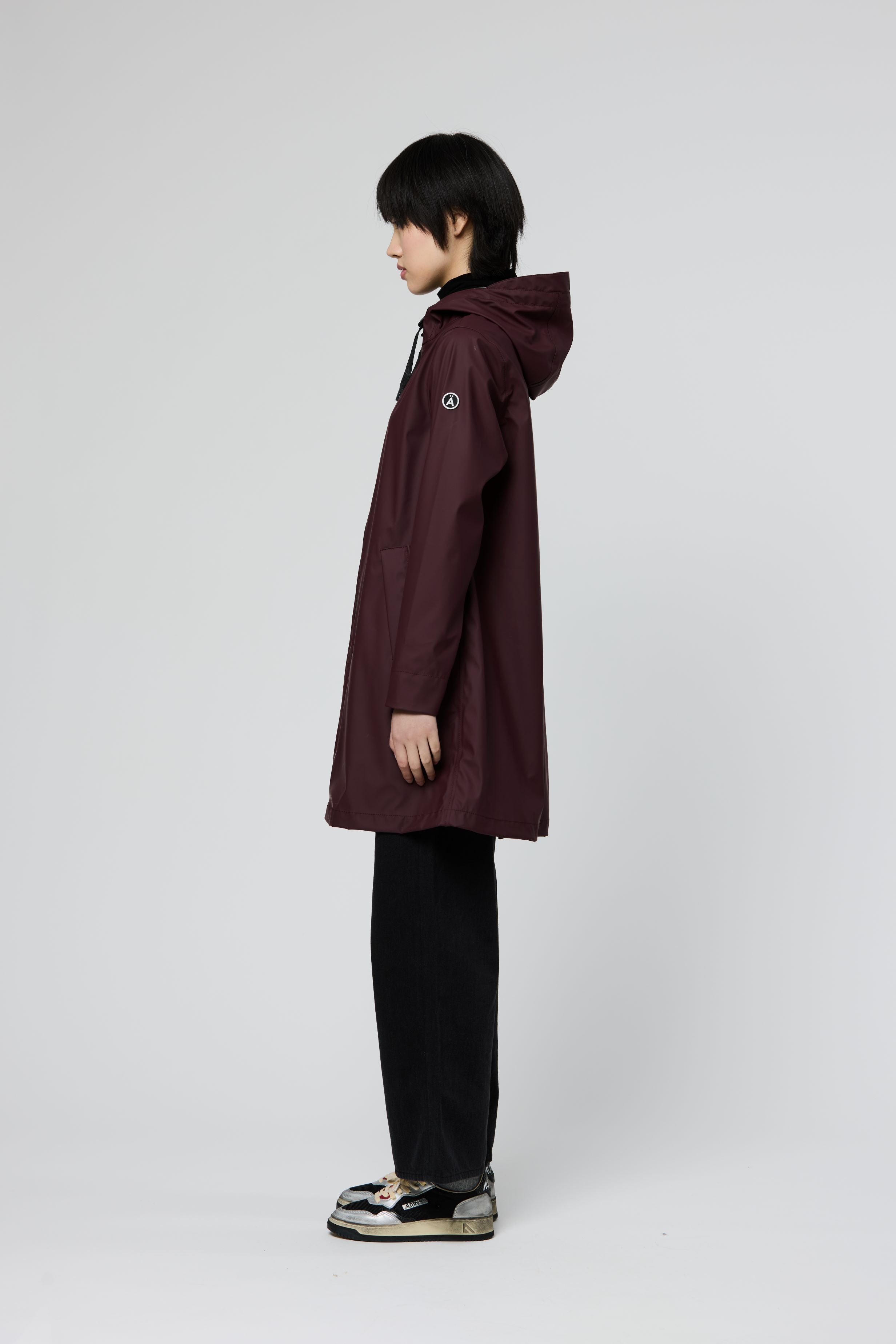 A model shown in profile wearing a long hooded raincoat in deep burgundy, paired with black trousers and black-and-white sneakers, against a light gray studio background.