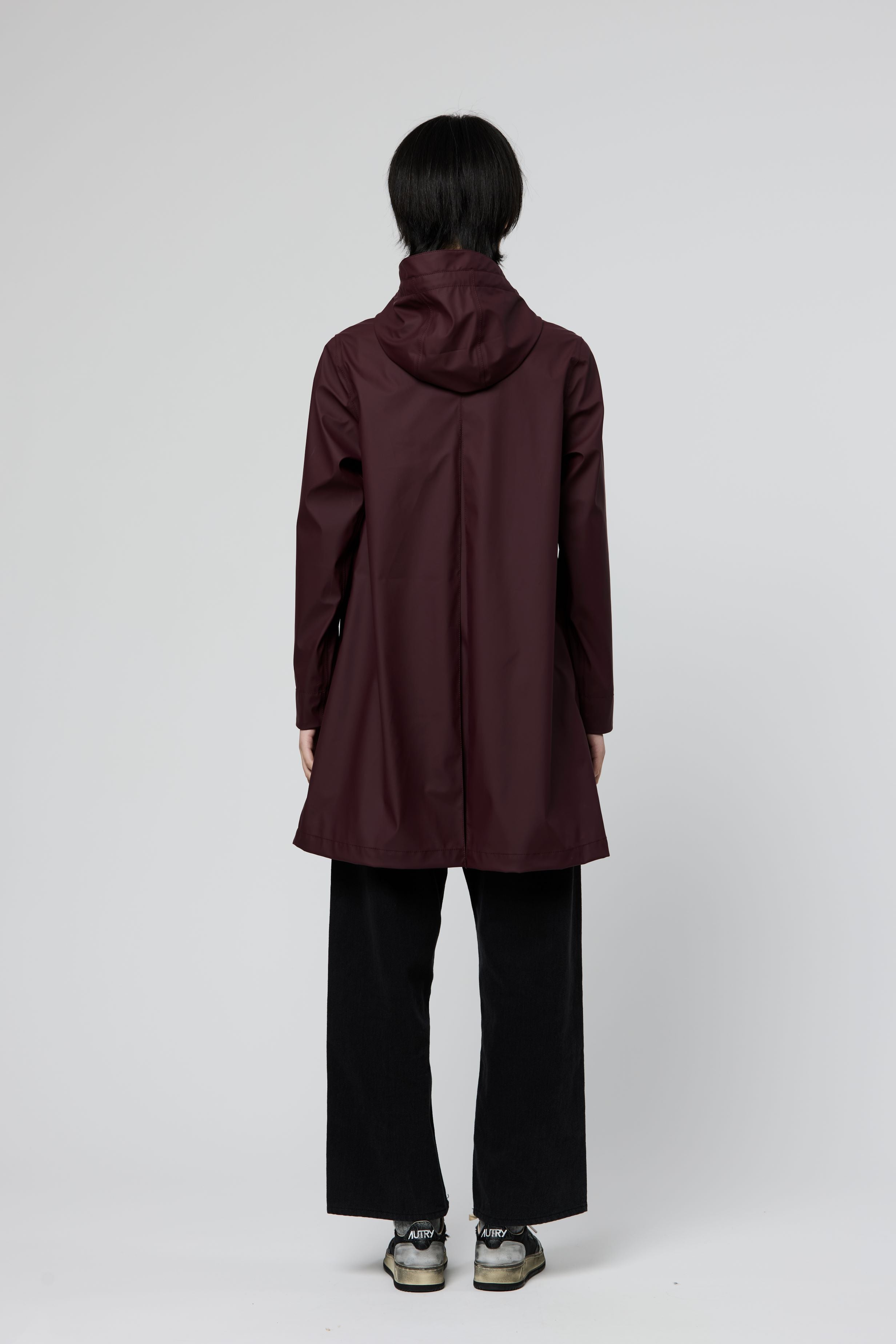 A person shown from the back wearing a hooded longline coat in deep burgundy, paired with black wide-leg pants and gray sneakers, against a light gray studio background.