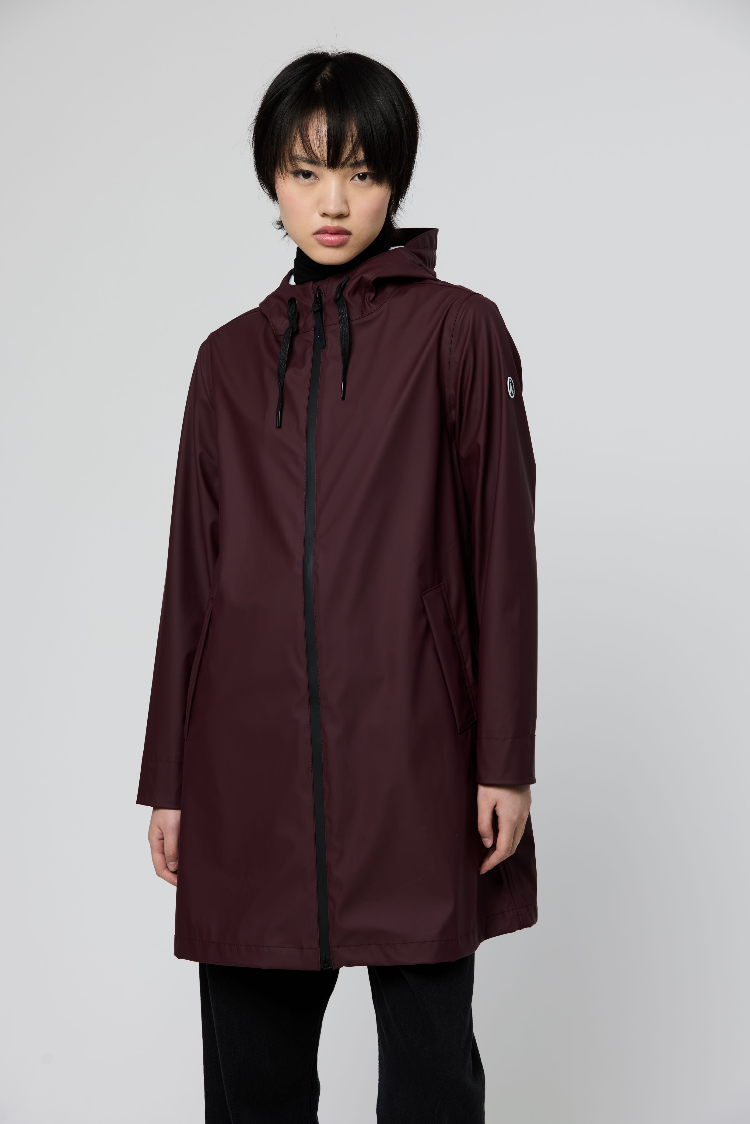A model wearing a long-sleeved hooded raincoat in a deep burgundy color, paired with black pants, against a plain light gray studio background.