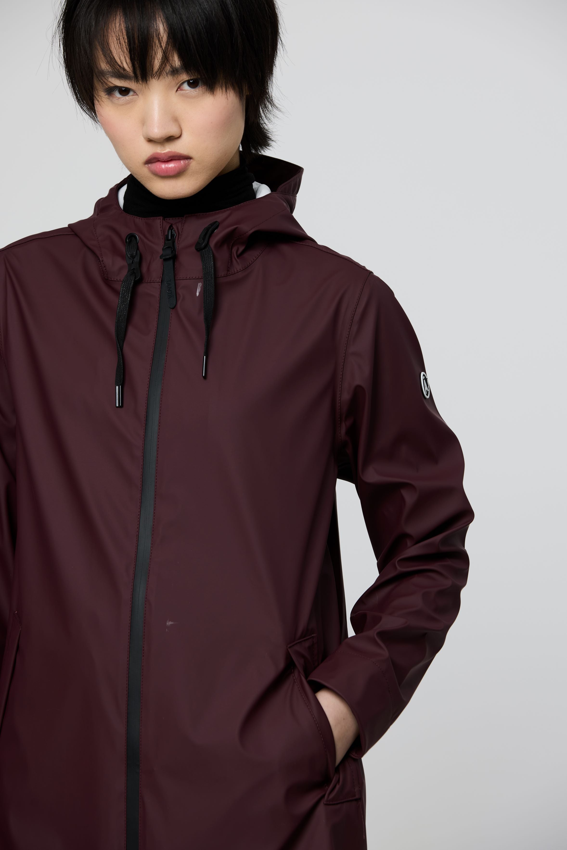 A model wearing a dark burgundy hooded rain jacket with black zippers and drawstrings, shown from the chest up against a light gray studio background.