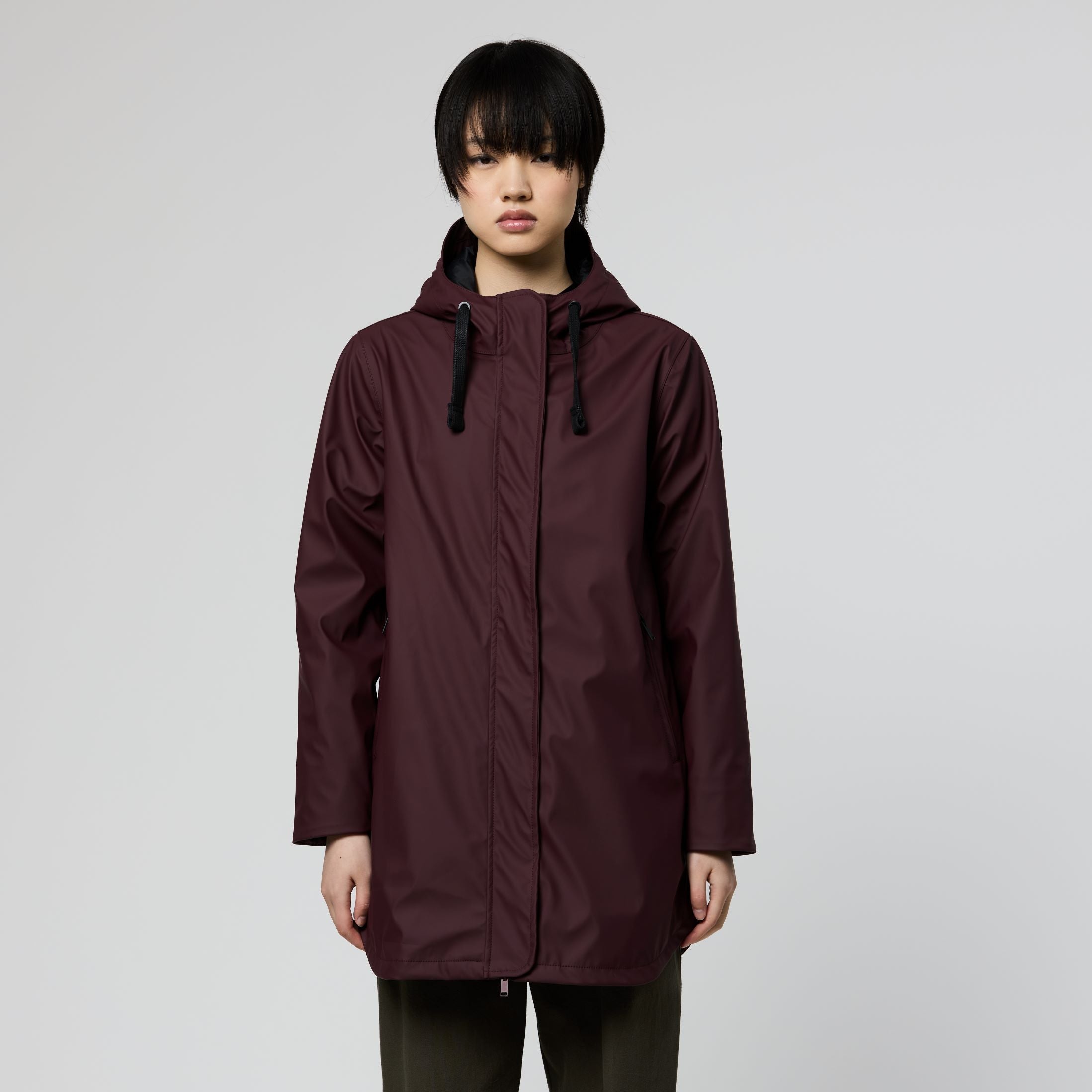 A model wearing a dark burgundy hooded raincoat with black drawstrings, paired with dark olive pants and brown shoes, standing against a light gray studio background.