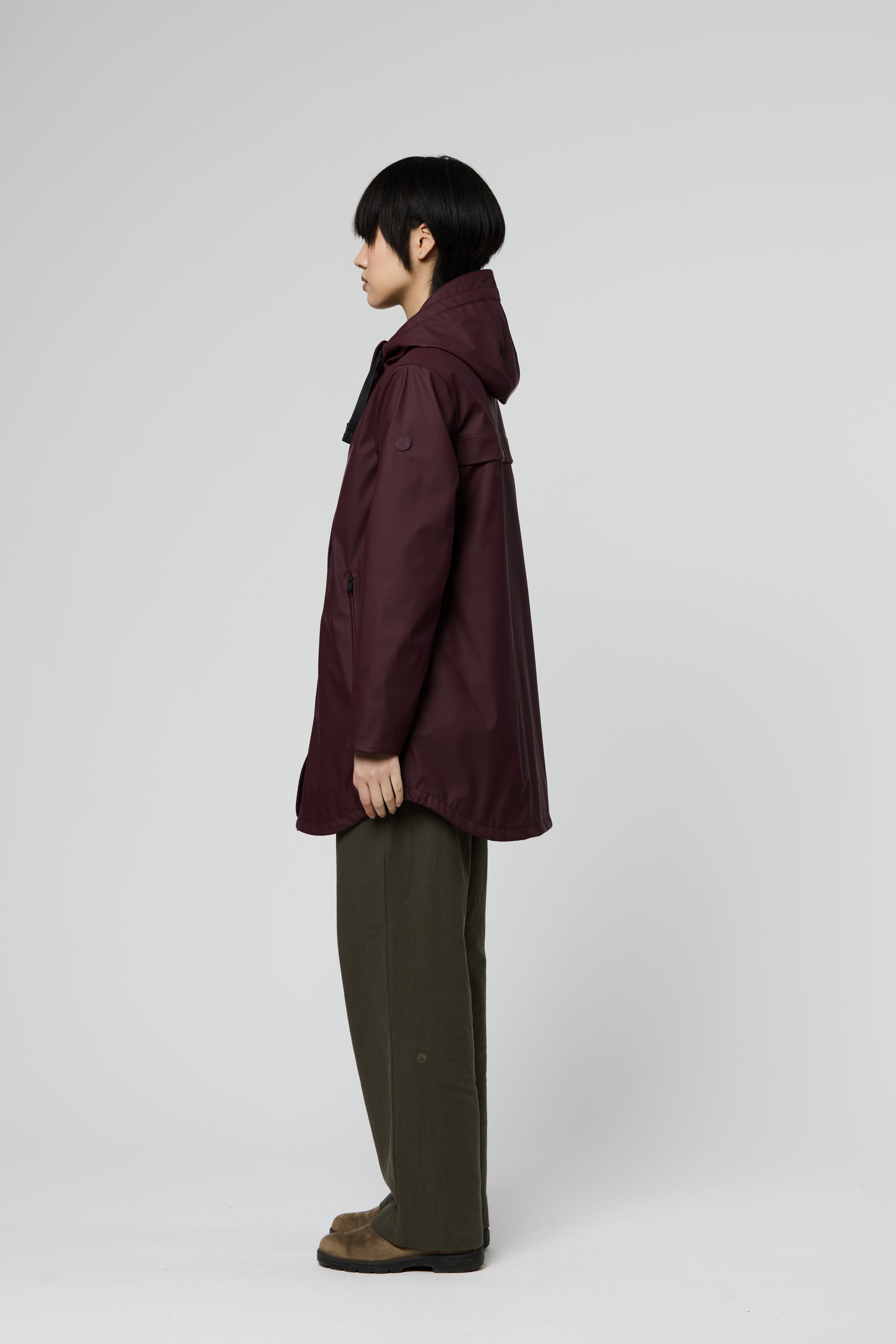 A model shown in profile wearing a dark plum hooded jacket, olive green wide-leg trousers, and brown shoes against a light gray studio background.