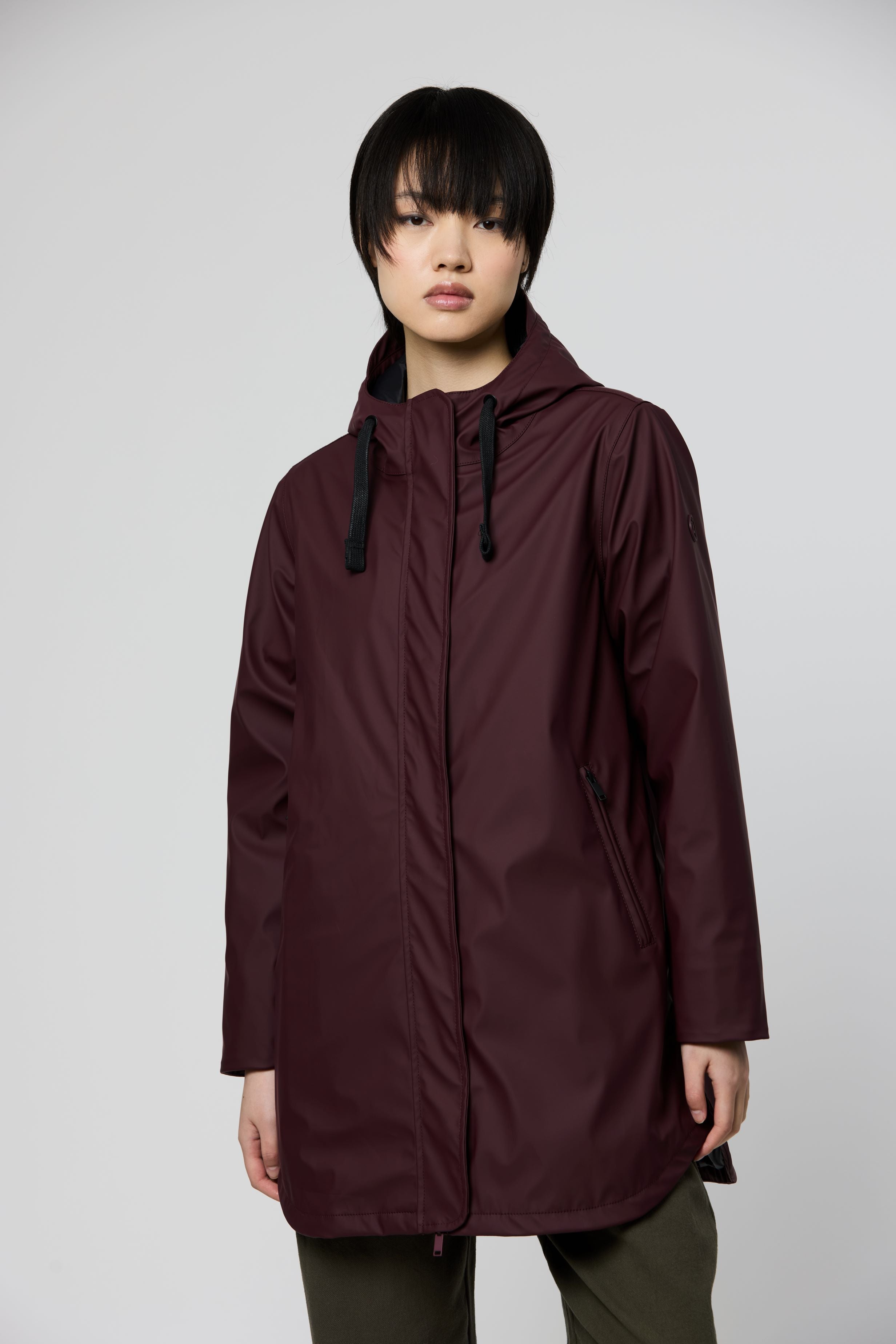 A model wearing a dark plum-colored hooded raincoat with black drawstrings, paired with olive green pants, against a solid light gray background.