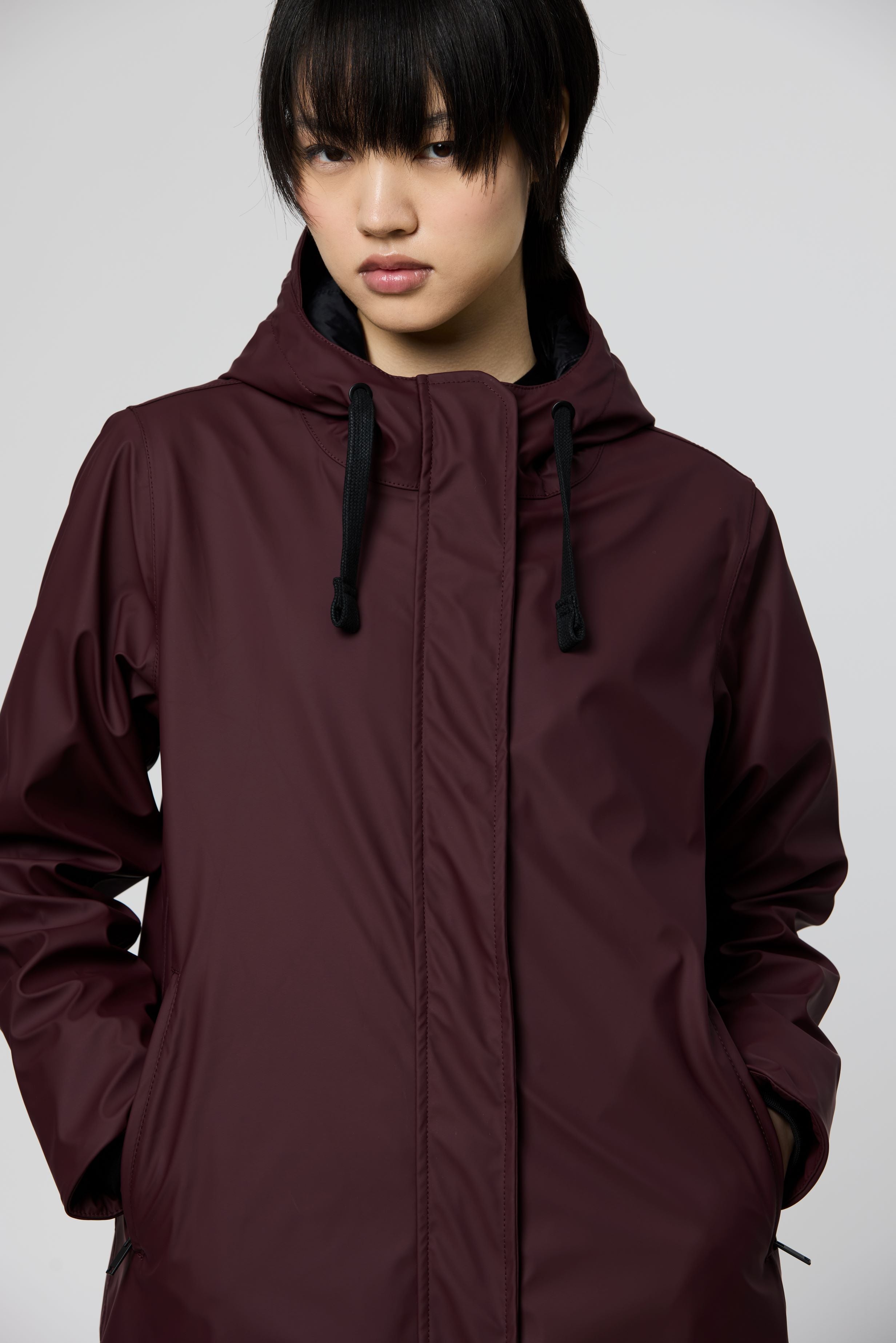 A model wearing a dark plum-colored hooded jacket with black drawstrings, photographed against a neutral light gray background.