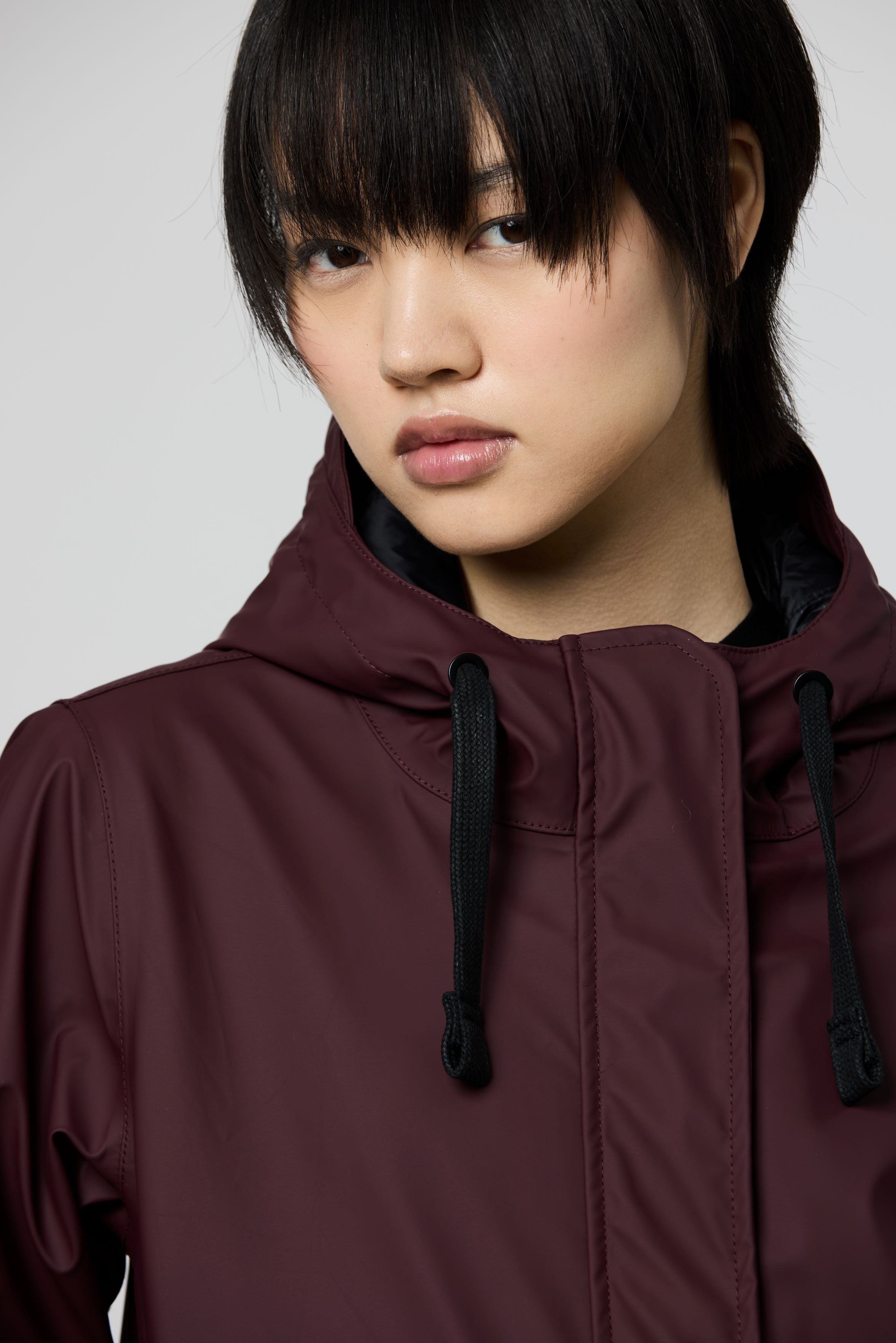 A close-up shot of a model wearing a dark plum-colored hooded jacket against a light gray background.
