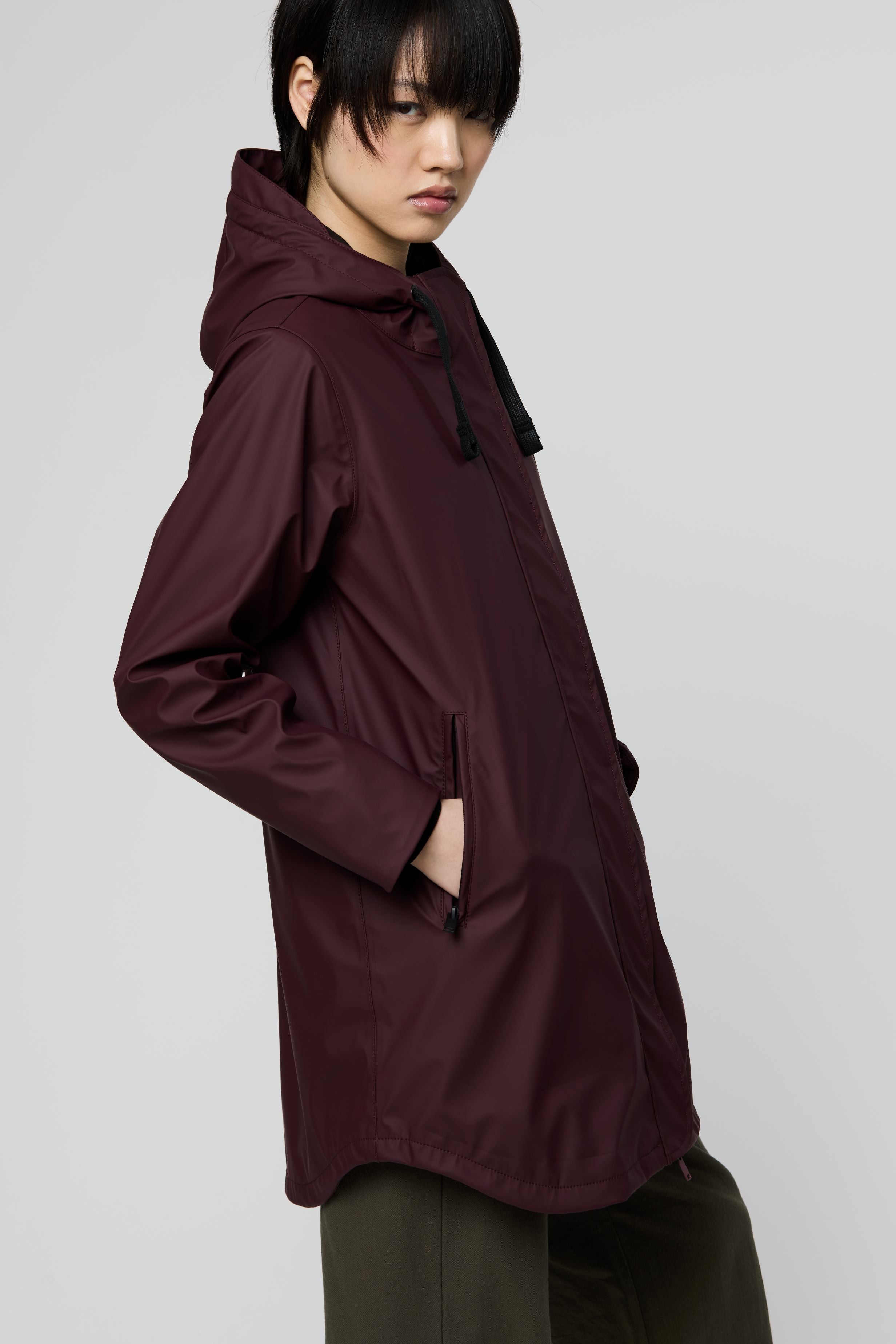 A model wearing a dark plum-colored hooded raincoat, shown from a side profile against a neutral light gray background.