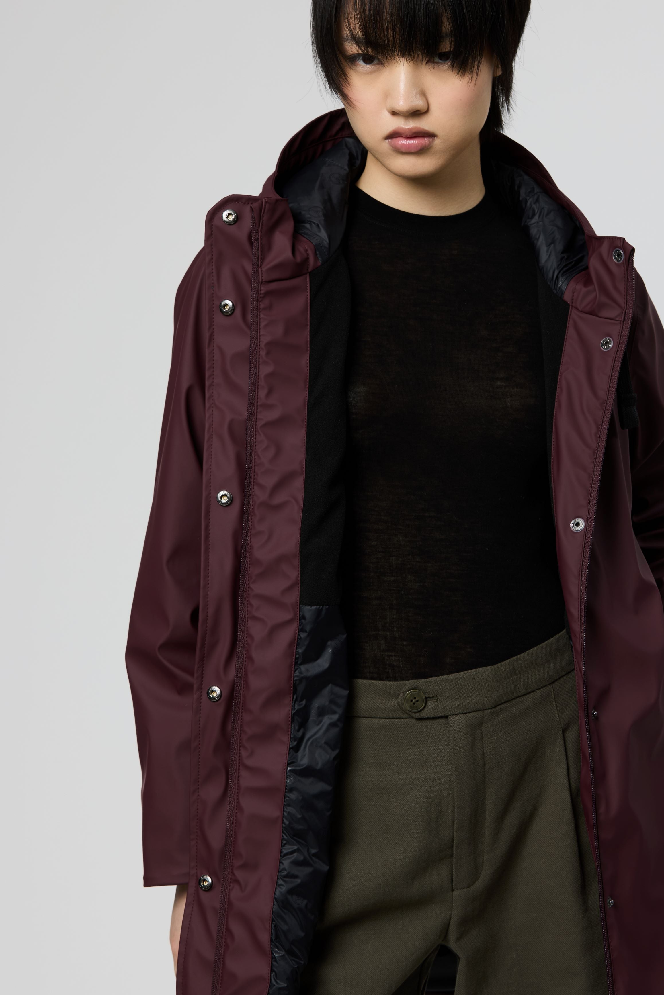 A model wearing a longline hooded raincoat in deep burgundy, styled over a black knit top and olive green trousers, photographed against a neutral light gray background.