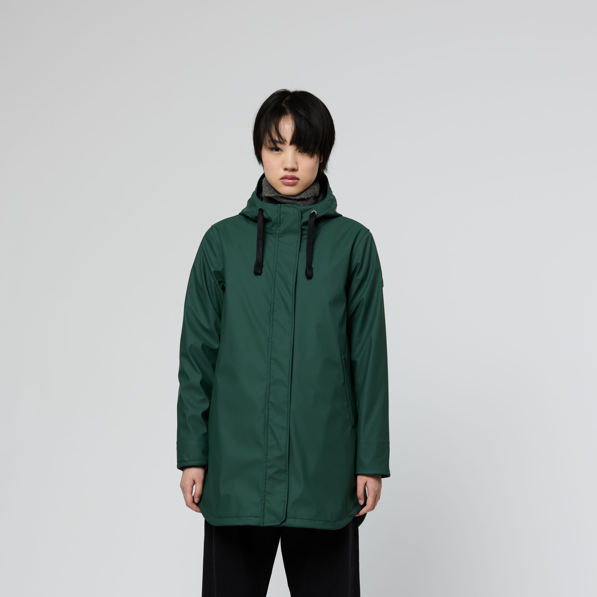 A model stands against a light gray studio background wearing a dark green hooded raincoat, black wide-leg pants, and black-and-white sneakers.