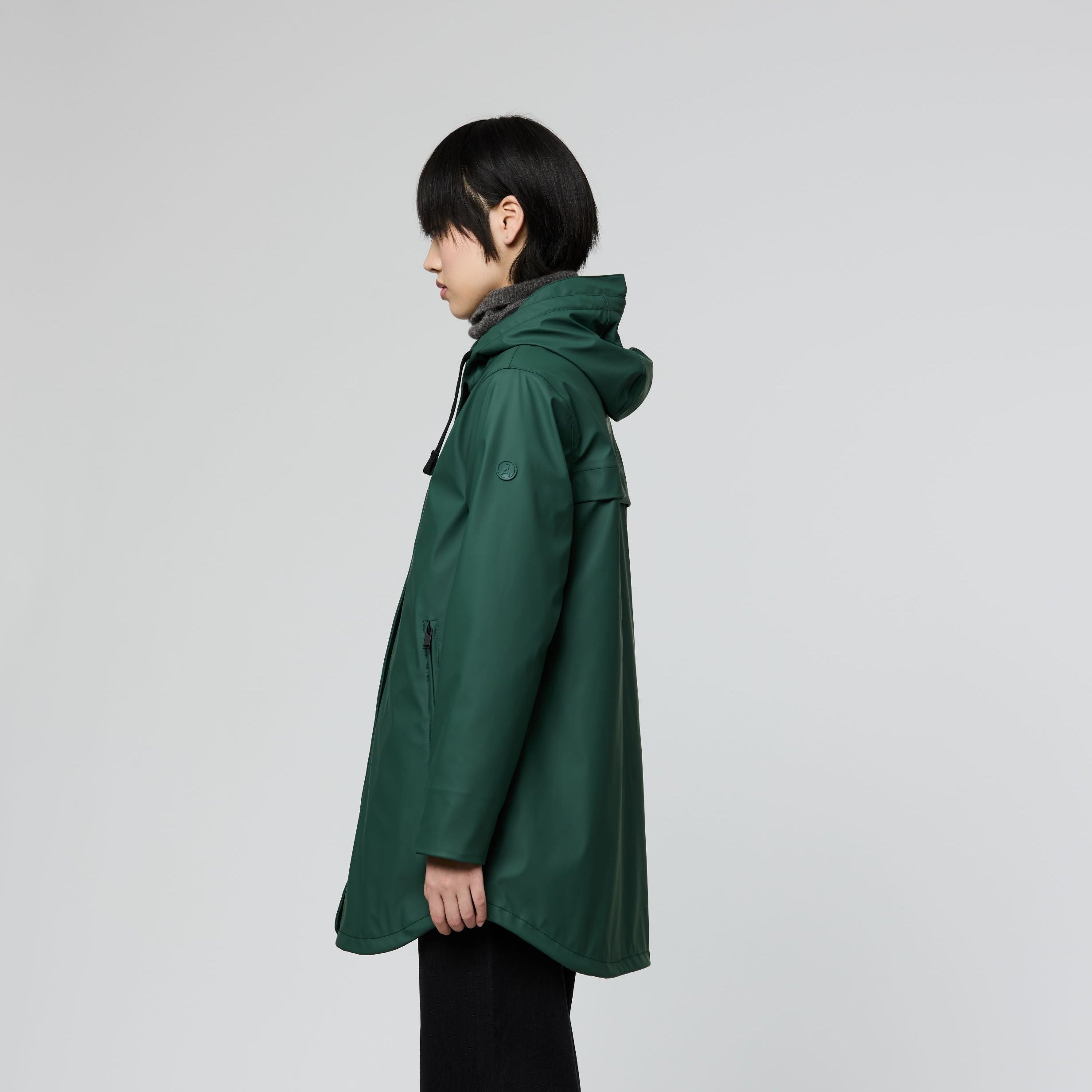 A child model shown in profile wearing a dark green hooded raincoat, black pants, and sneakers against a light gray studio background.