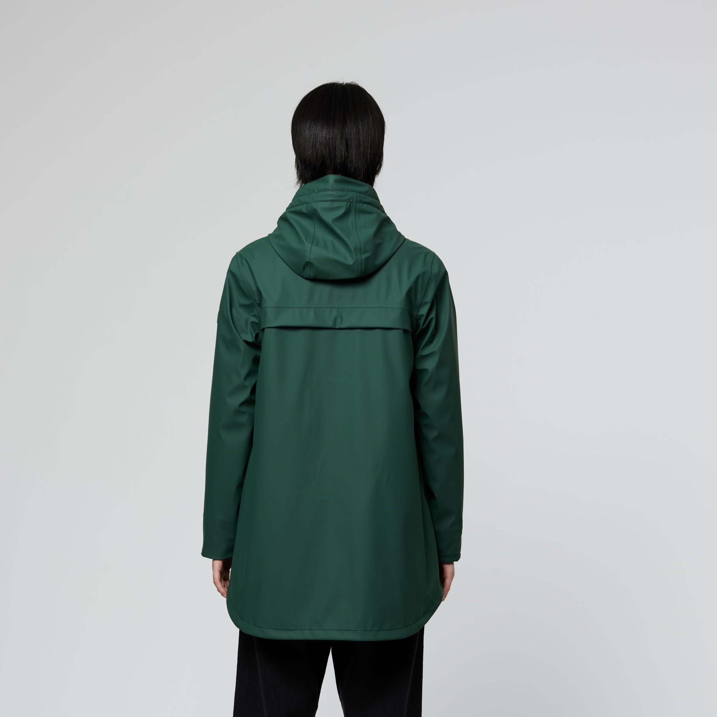 A person shown from the back wearing a dark green hooded jacket, black wide-leg pants, and gray sneakers against a light gray studio background.