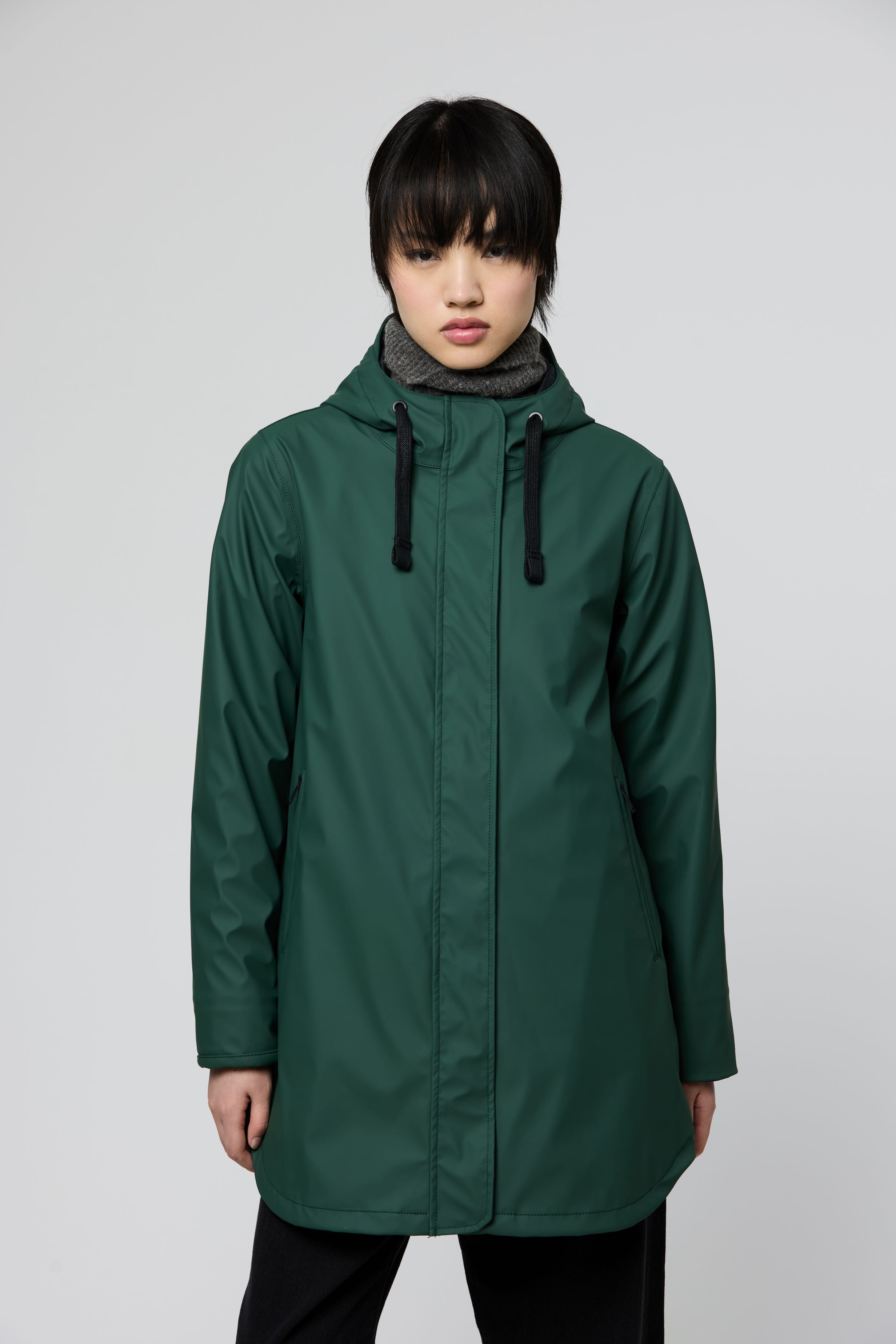 A model wearing a dark green hooded raincoat with black drawstrings, photographed against a neutral light gray studio background.