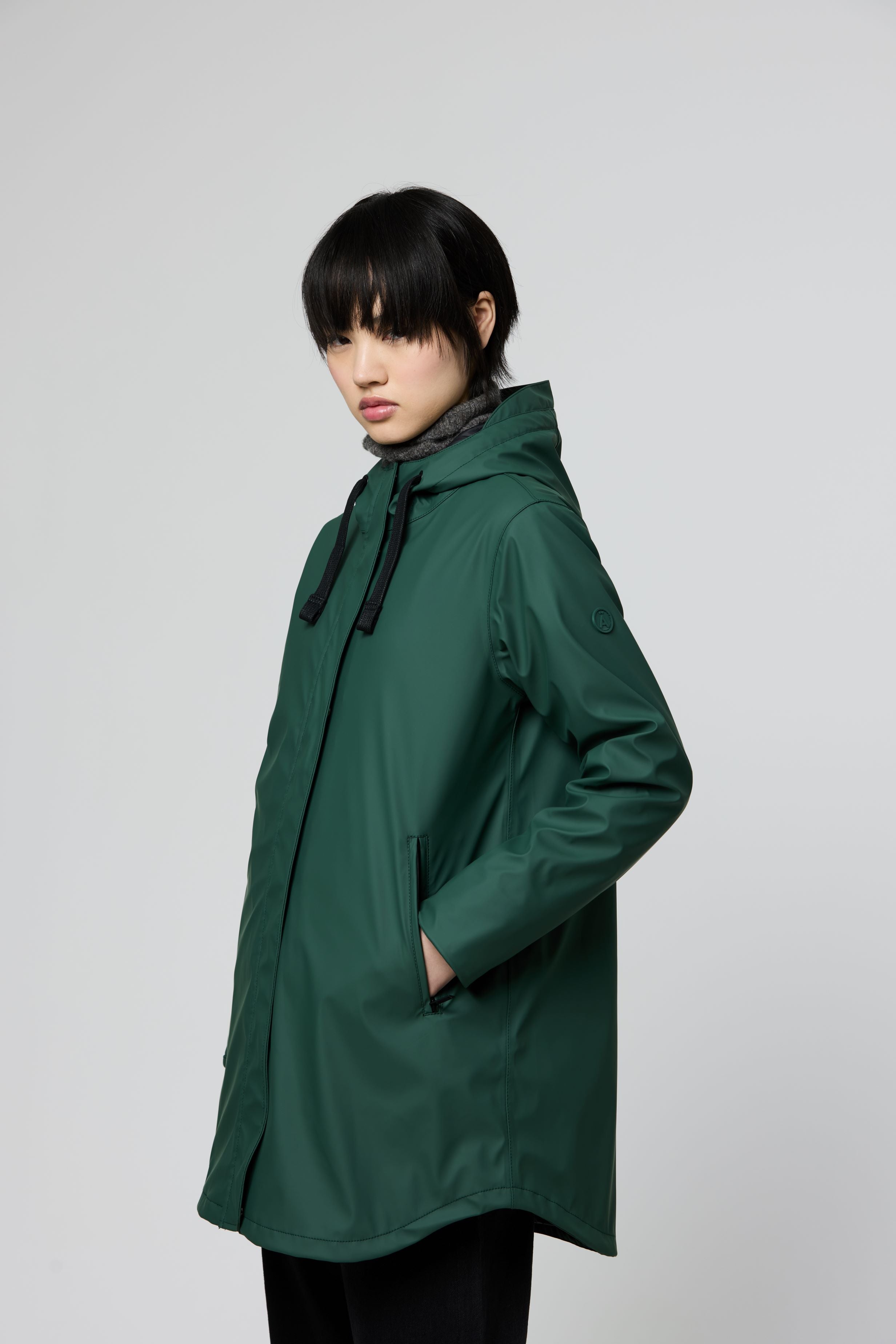 A model wearing a dark green hooded raincoat with black drawstrings, standing against a light gray studio background.