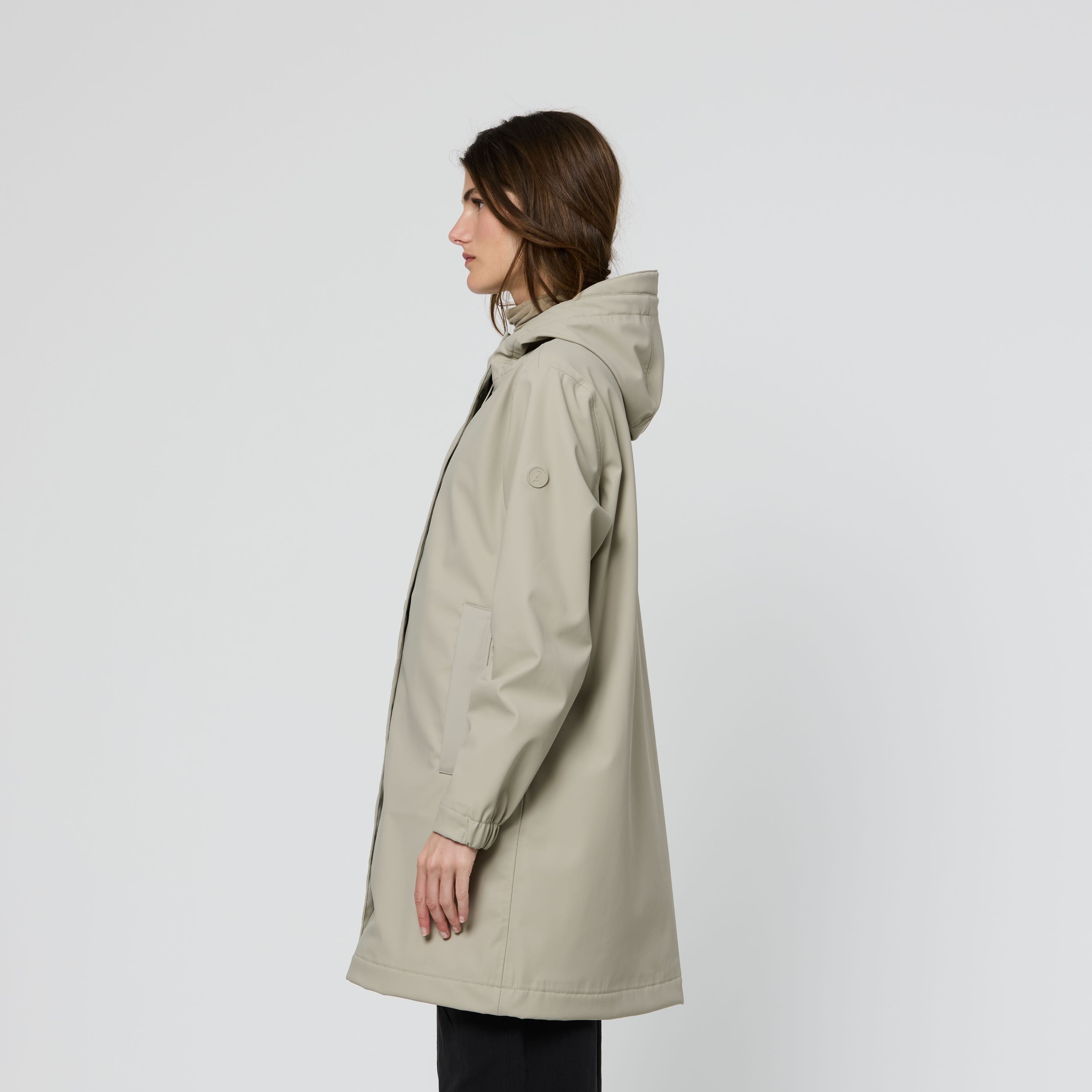 A woman shown in profile wearing a light beige hooded raincoat, black pants, and brown sneakers against a plain light gray studio background.
