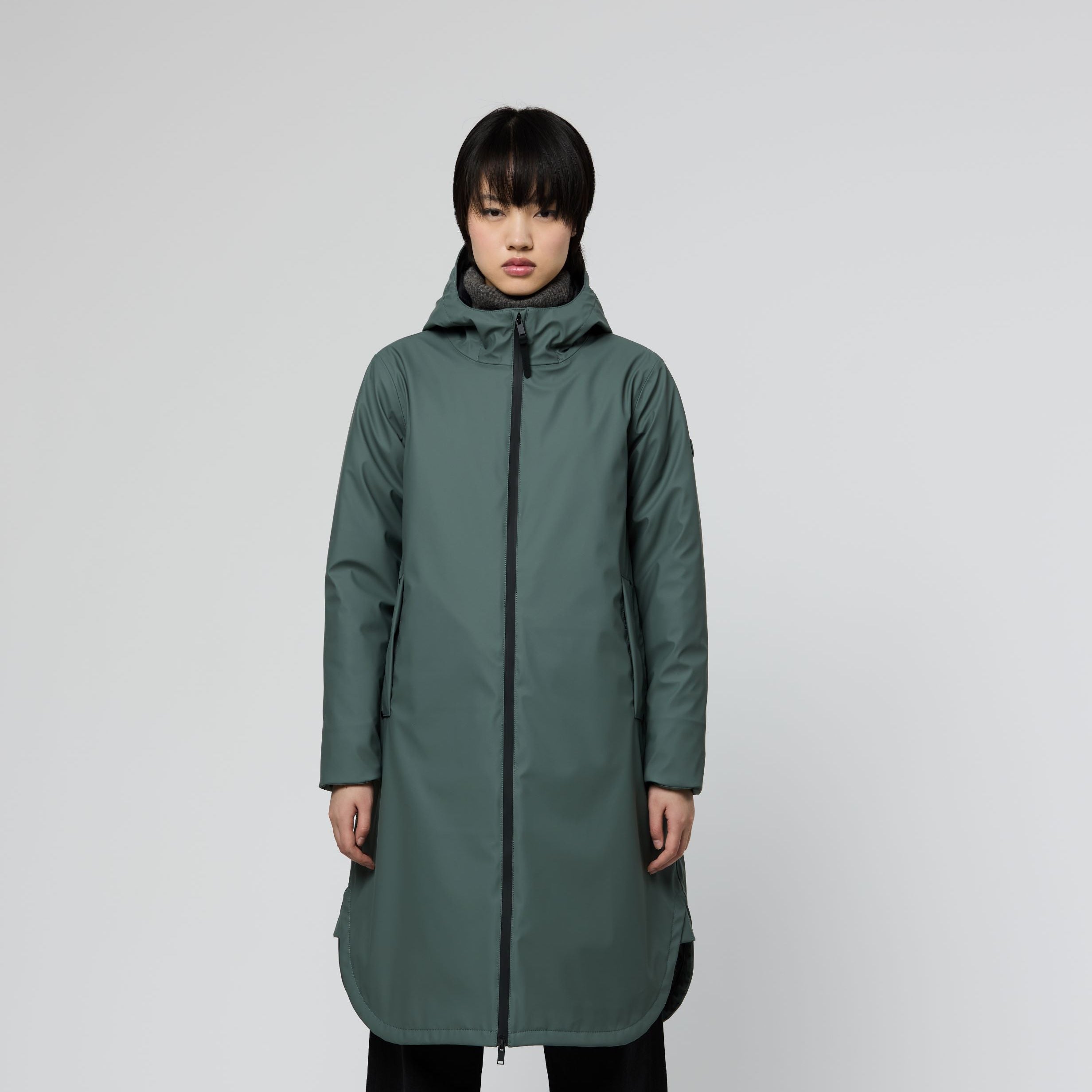 A model wearing a long, dark green hooded puffer coat with black wide-leg pants and sneakers, standing against a light gray studio background.