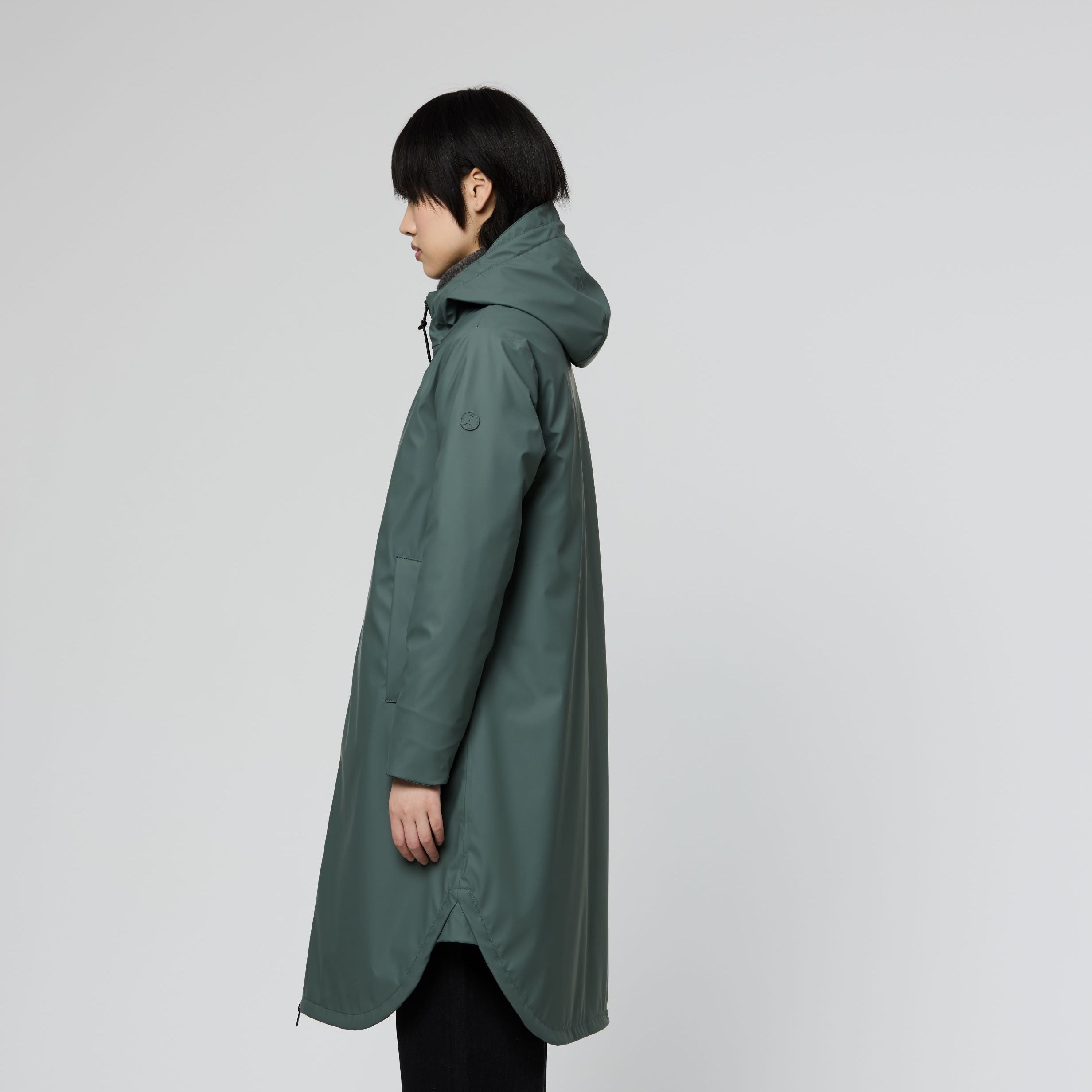 A model shown in profile wearing a long, dark green hooded raincoat, black pants, and sneakers against a plain light gray studio background.