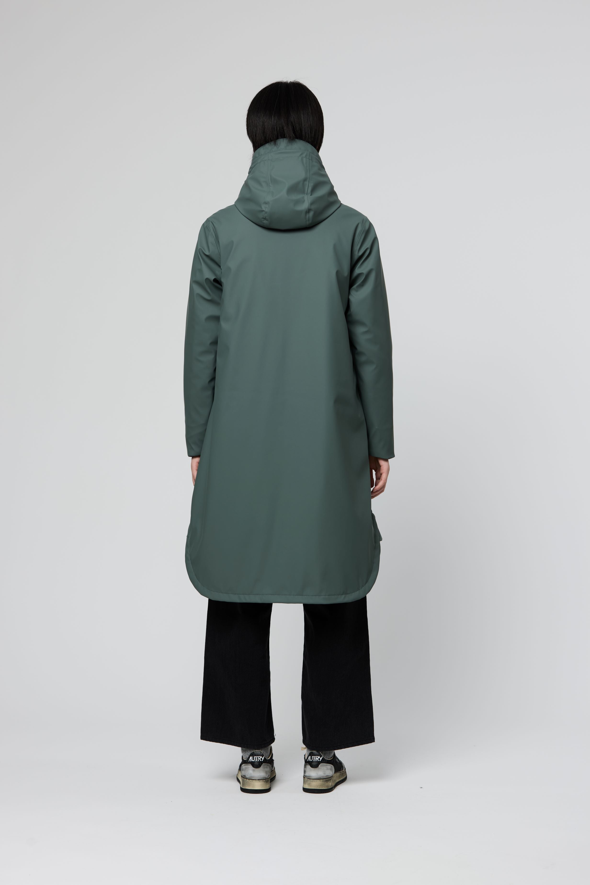 A person shown from the back wearing a long hooded coat in muted sage green, paired with black wide-leg pants and gray sneakers, against a plain light gray studio background.