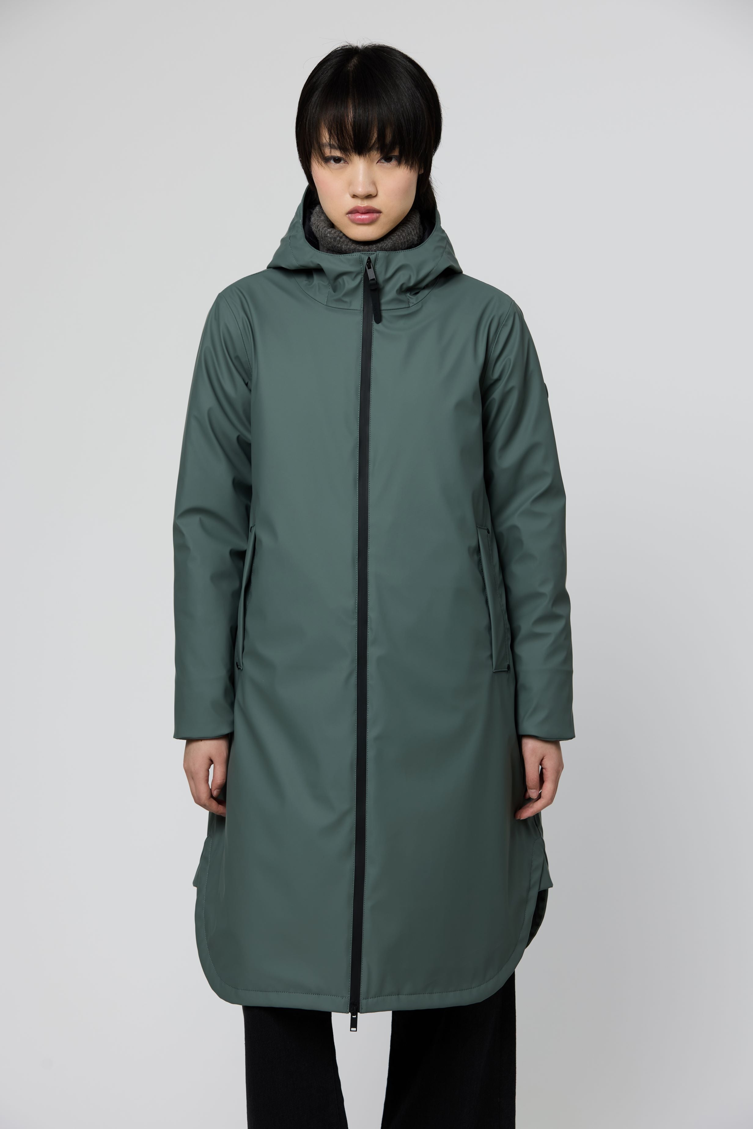 A model wearing a long, dark green hooded raincoat with a black front zipper, photographed against a neutral light gray studio background.